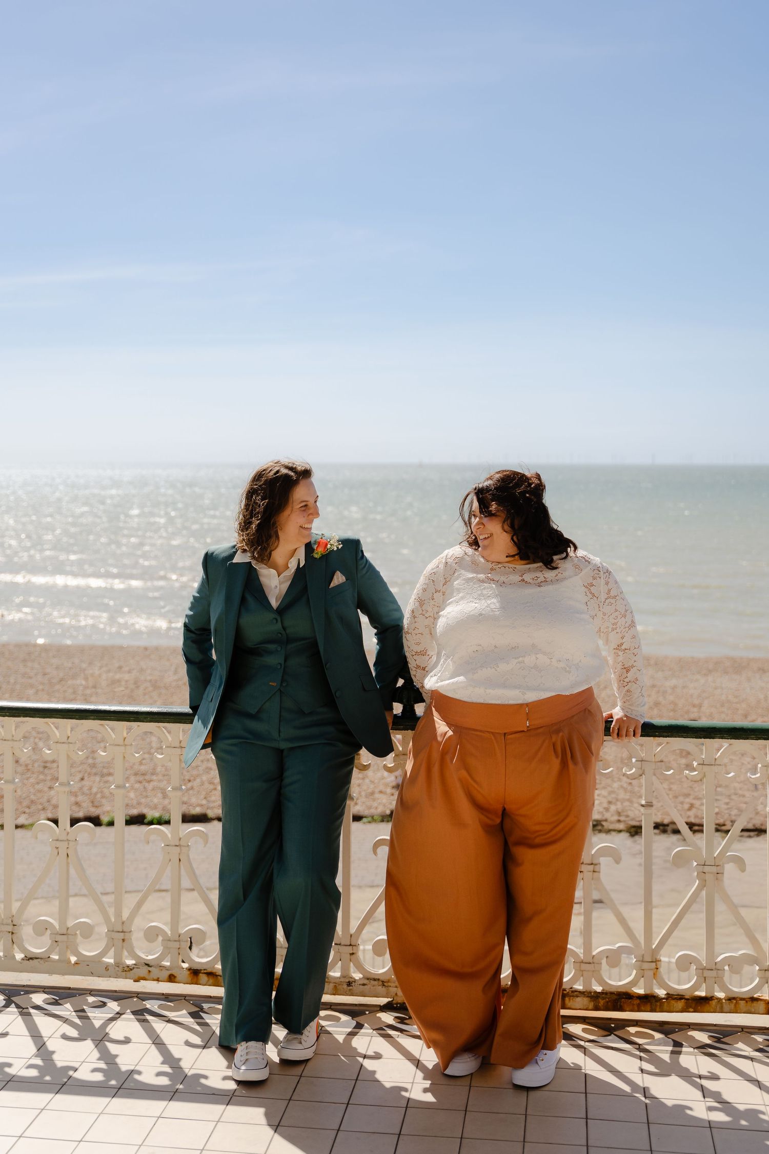Two women in elegant outfits stand on a seaside balcony, smiling at each other on a sunny day.