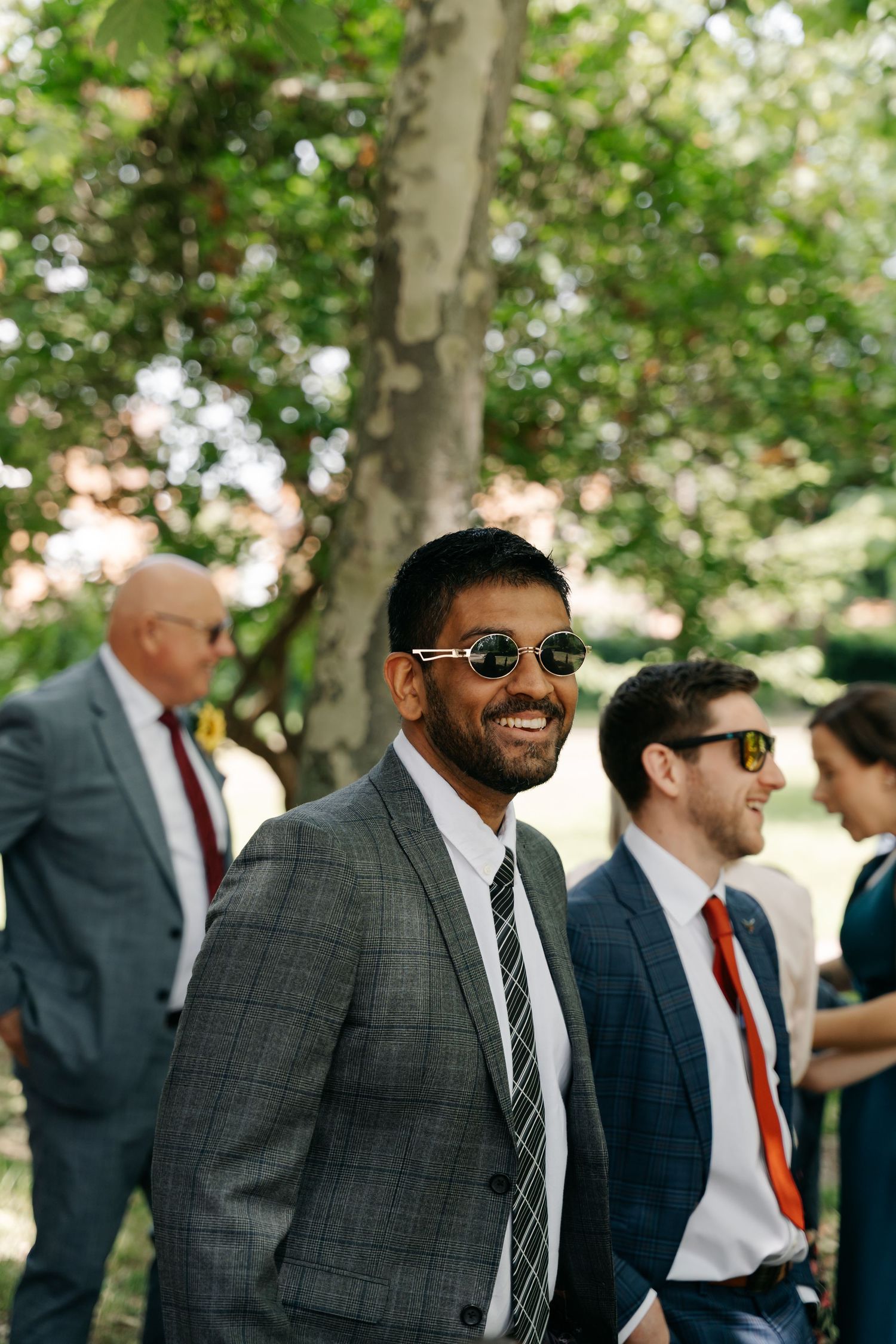 Smiling man in gray suit and round sunglasses stands outdoors among wedding guests beneath green trees.