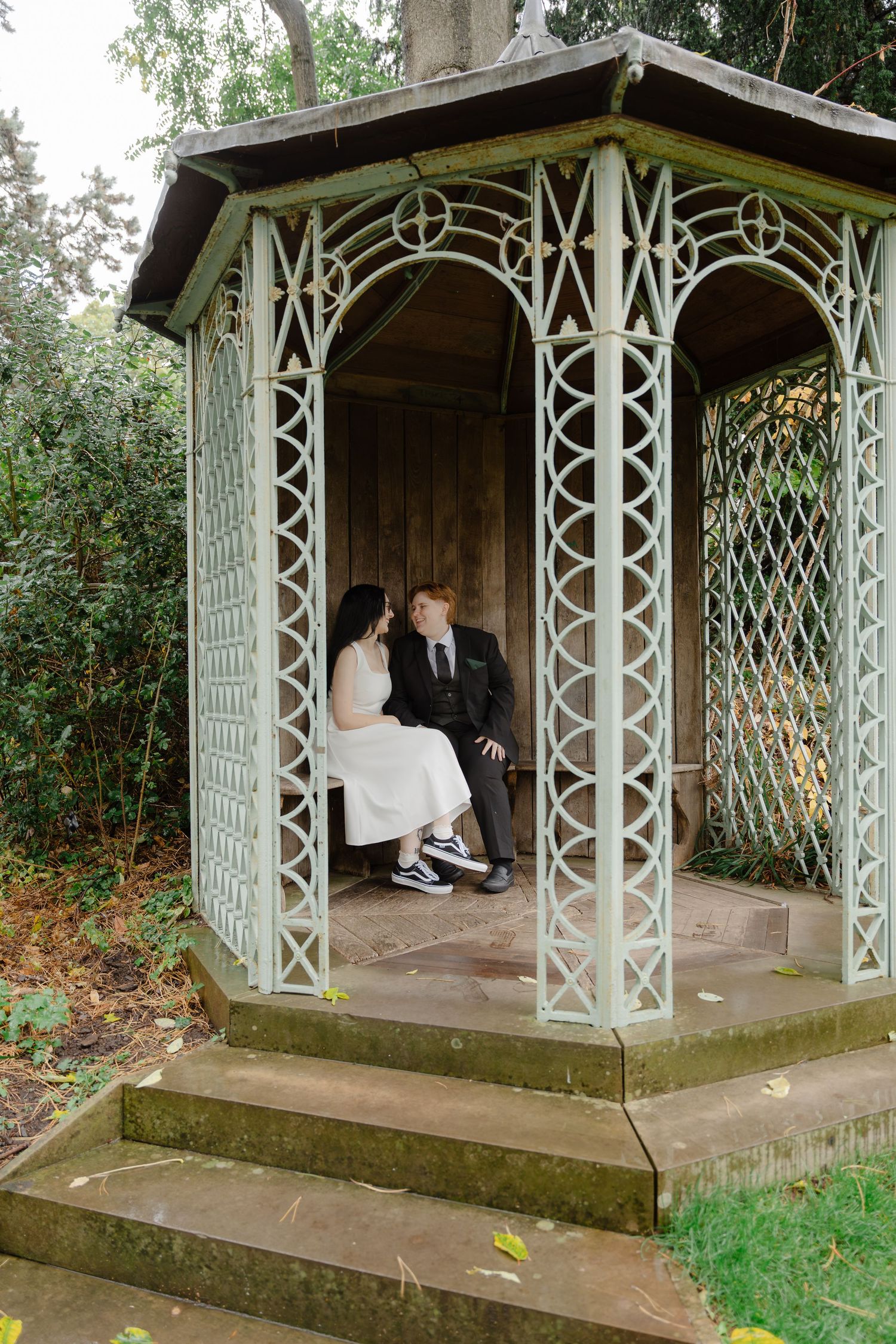A couple in wedding attire sits together in a vintage white lattice garden gazebo surrounded by lush greenery.