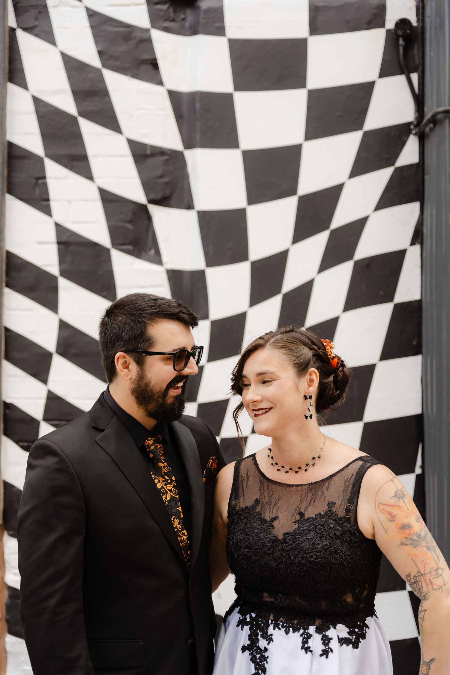 Couple smiling at each other in front of a black and white checkered wall, wearing elegant black outfits.