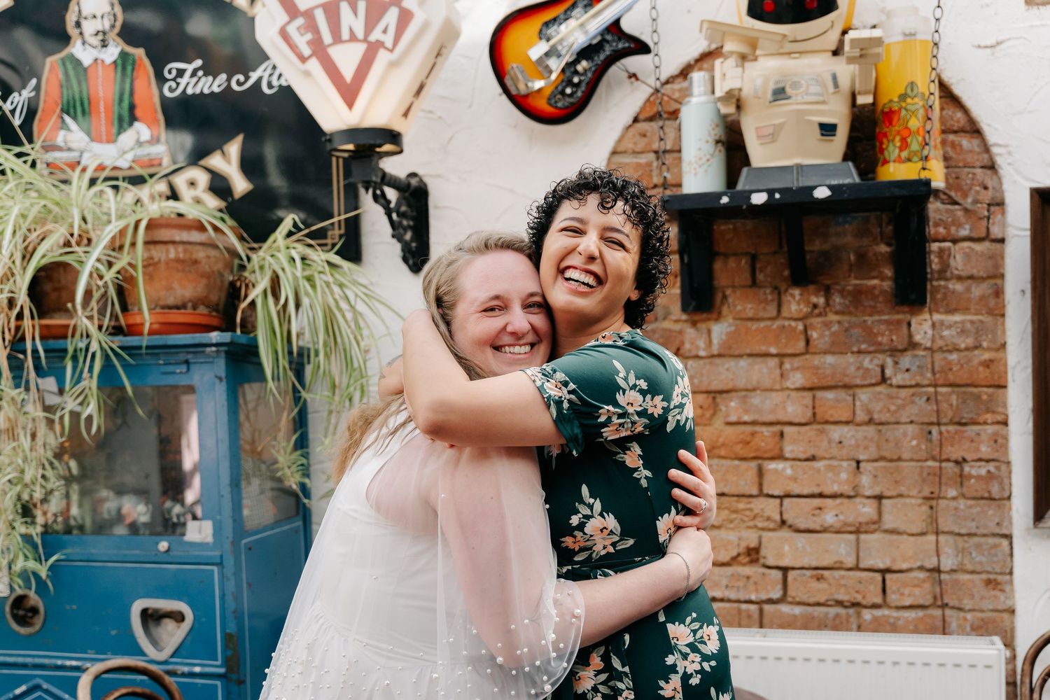 Two women hugging and smiling at a rustic wedding venue with brick walls and vintage decor.