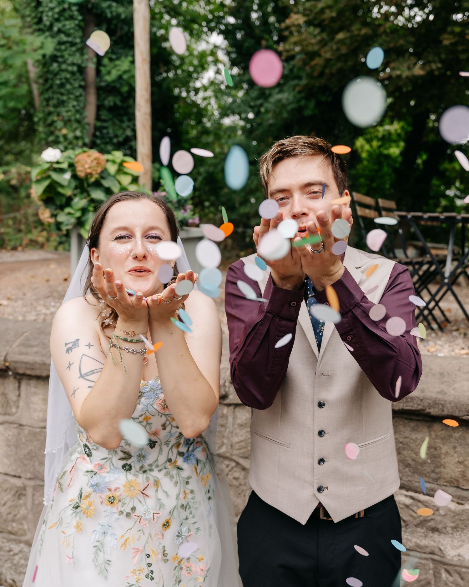 Bride and groom blowing colorful confetti at outdoor wedding celebration, bride wearing floral dress with veil.