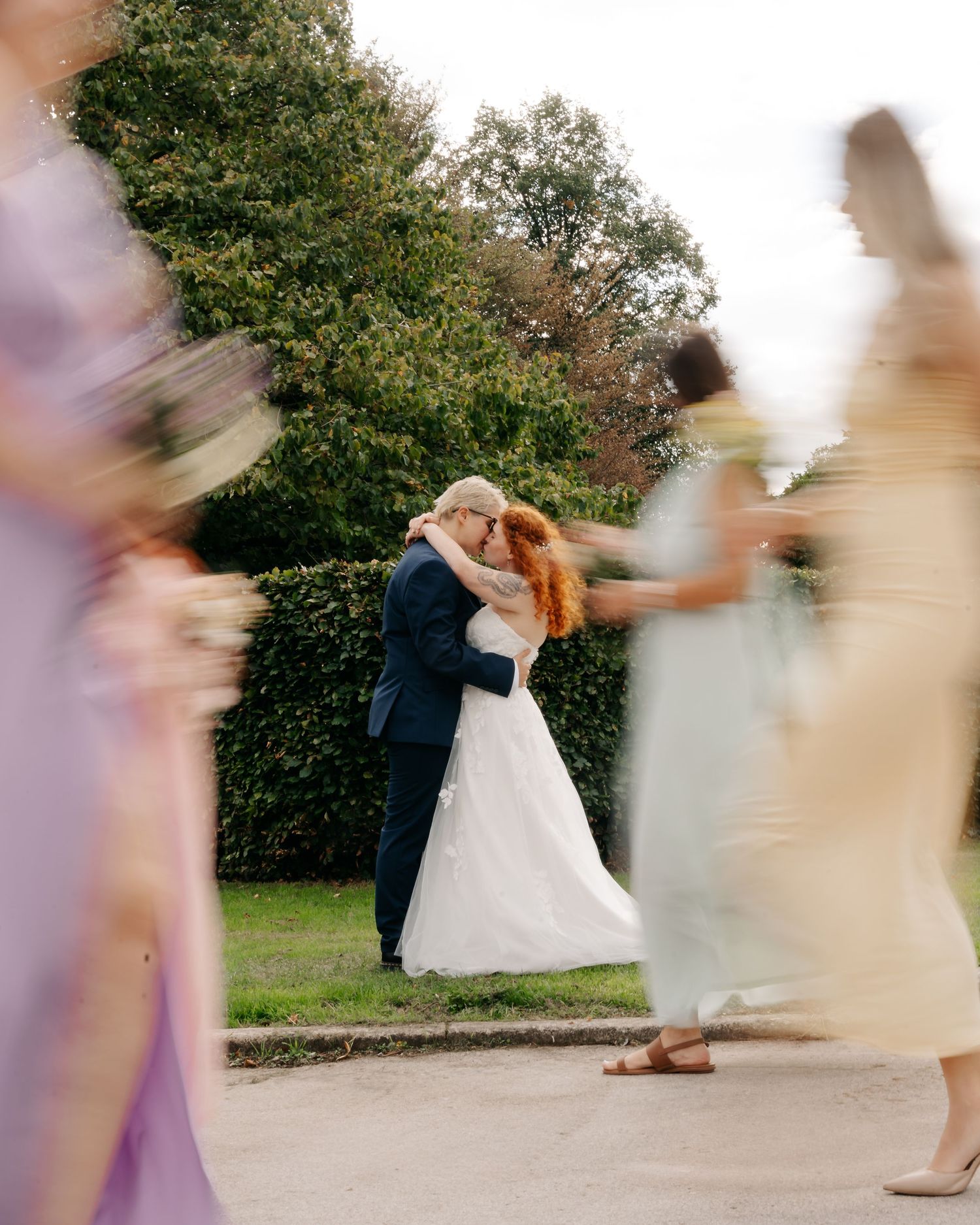 Bride and groom share a kiss surrounded by blurred bridesmaids in pastel dresses at an outdoor wedding.