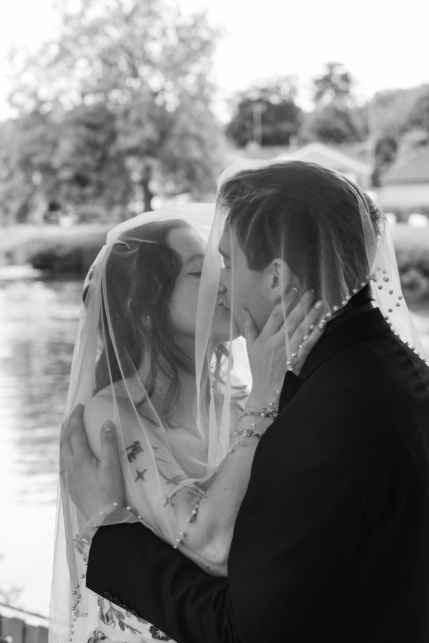 Black and white photo of a bride and groom sharing a kiss under a veil near a tranquil lake surrounded by trees.
