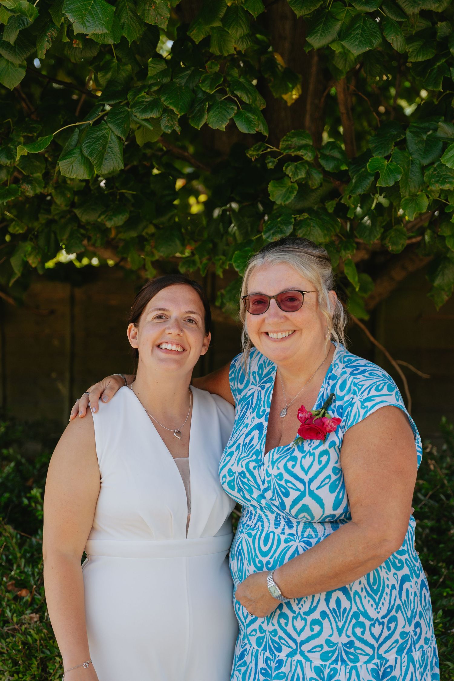 Two women smiling outdoors under green foliage, one in white dress, other in blue patterned top with red flower.