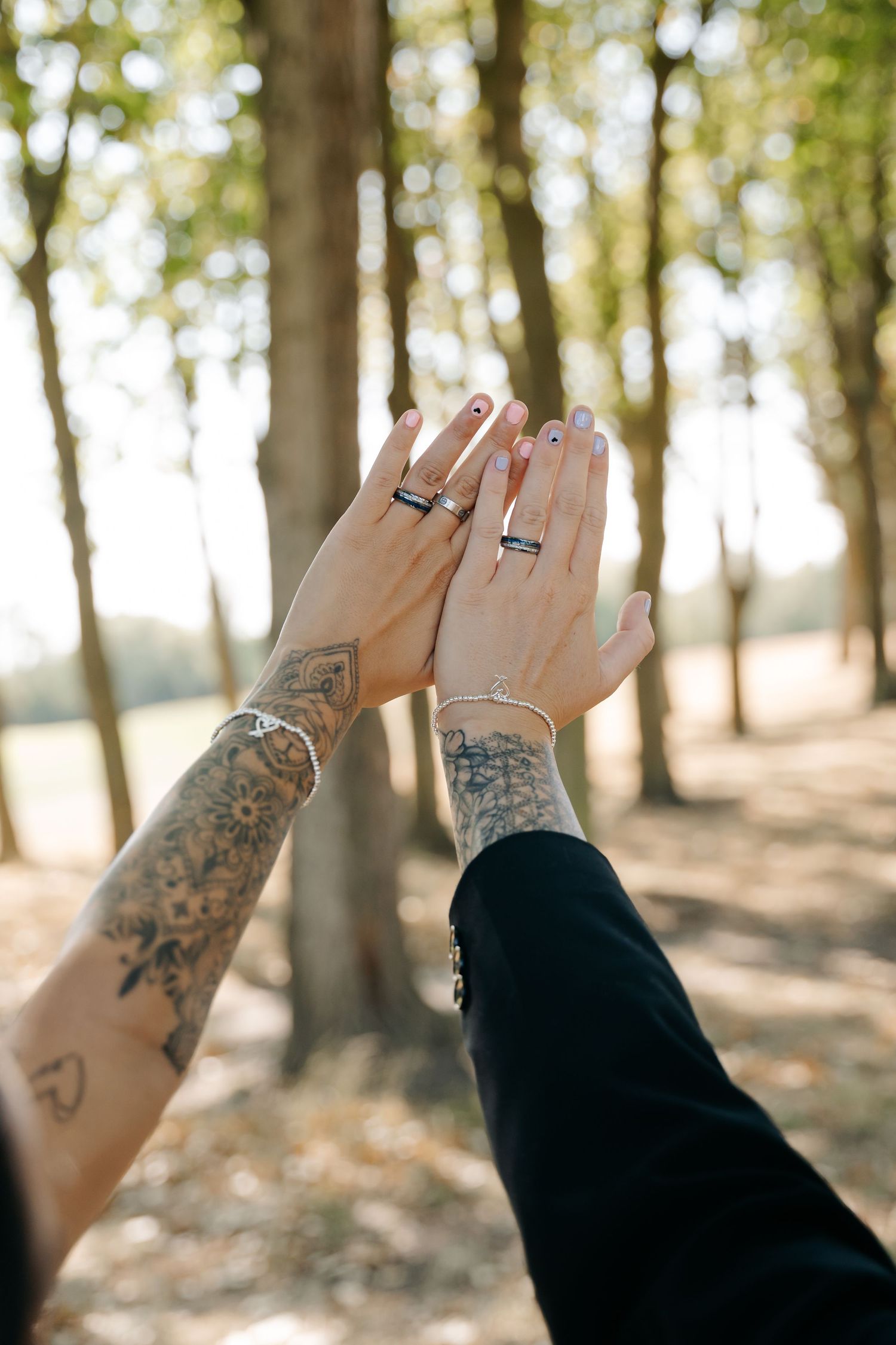 Two tattooed hands with matching rings touch in a sunlit forest, symbolizing love and commitment.