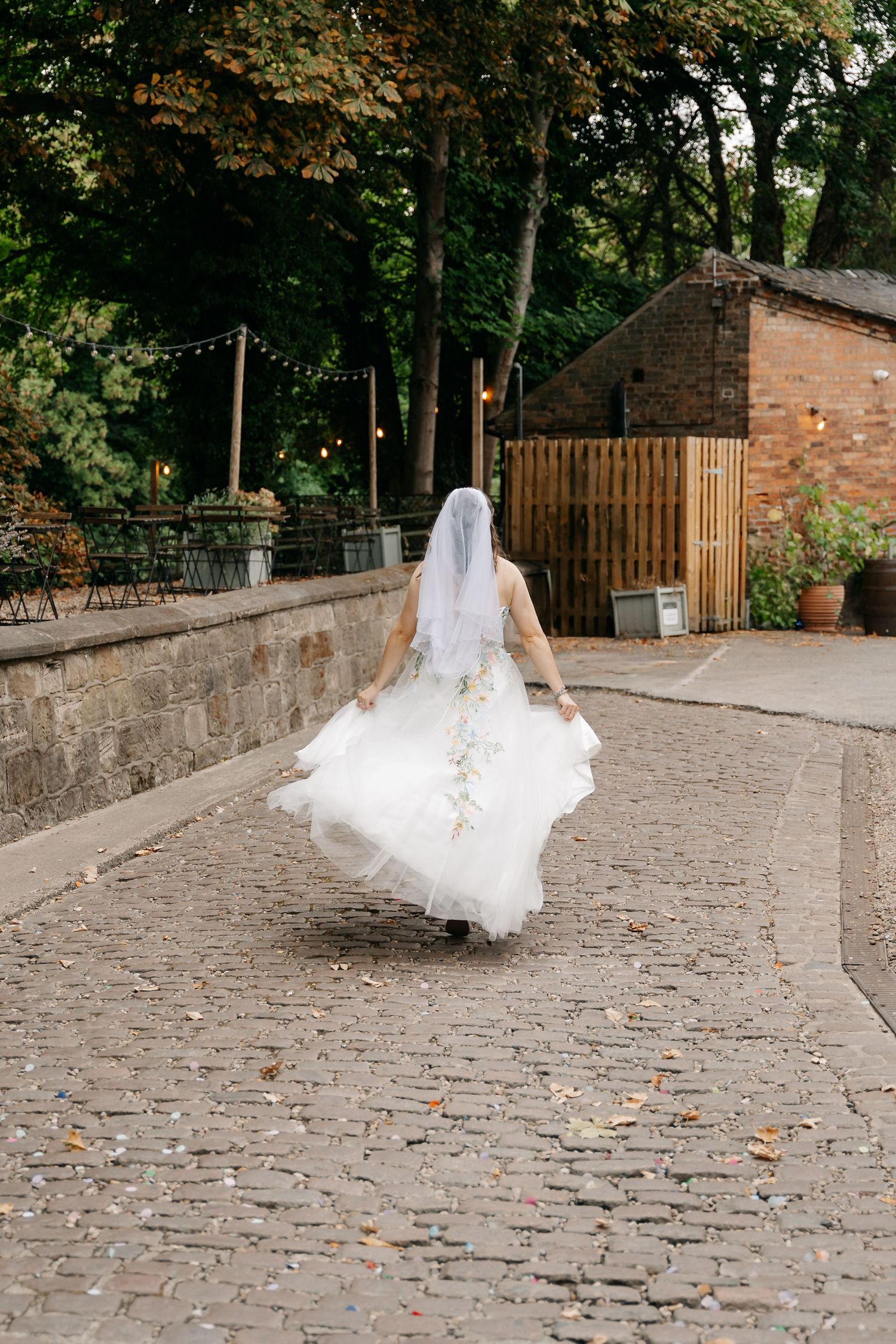 Bride in white gown and veil walks away on cobblestone path surrounded by lush trees and string lights at outdoor venue.