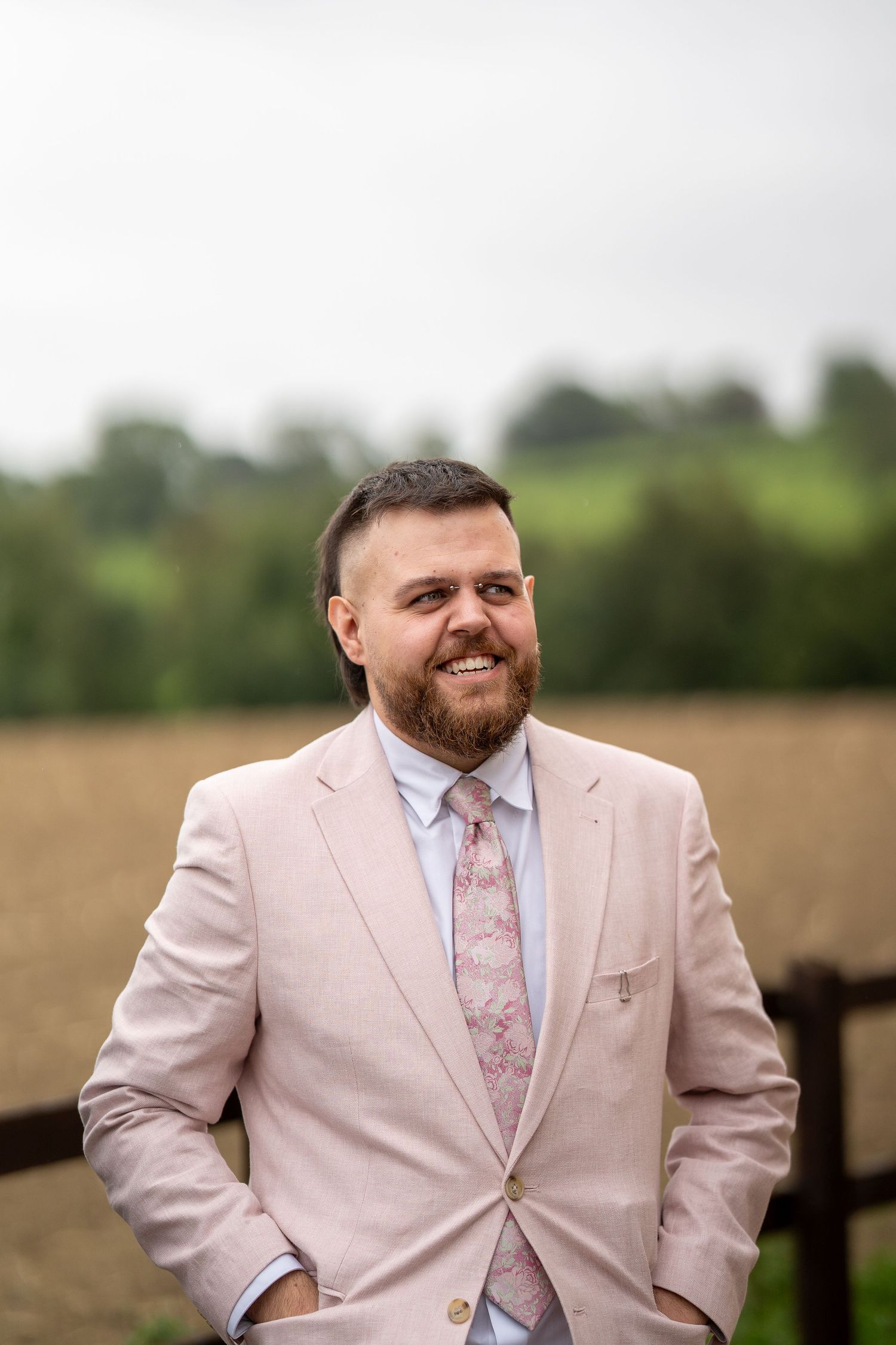 Smiling bearded man in a pink suit and floral tie stands outdoors with green fields in the background.