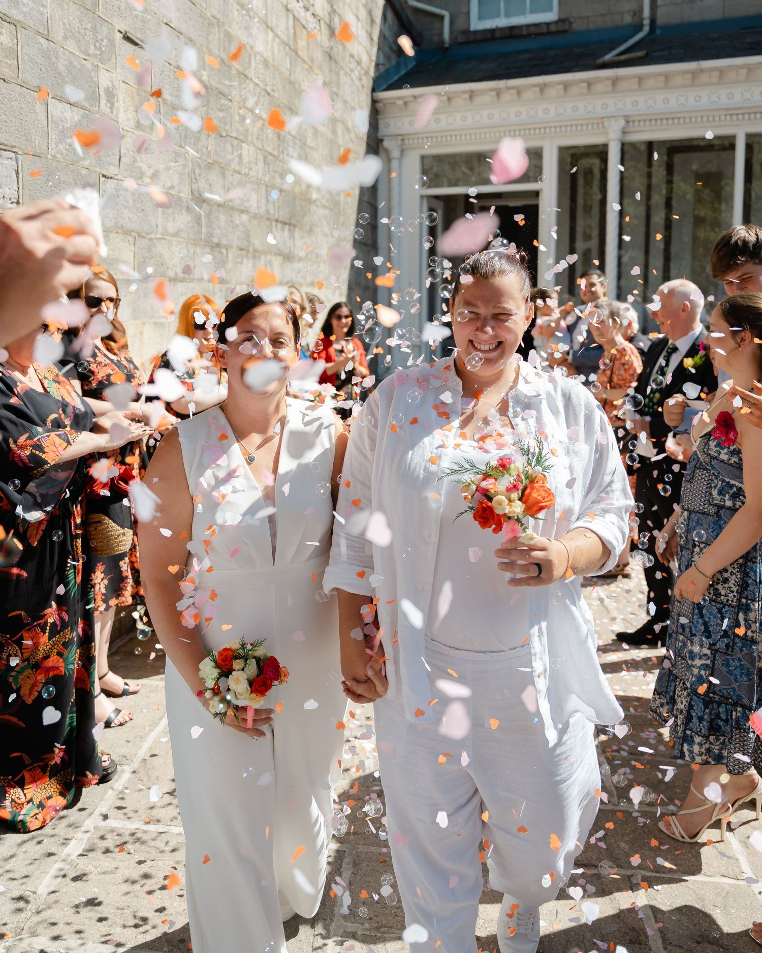 Two brides in white walk through confetti shower after wedding ceremony, guests celebrating outdoors.