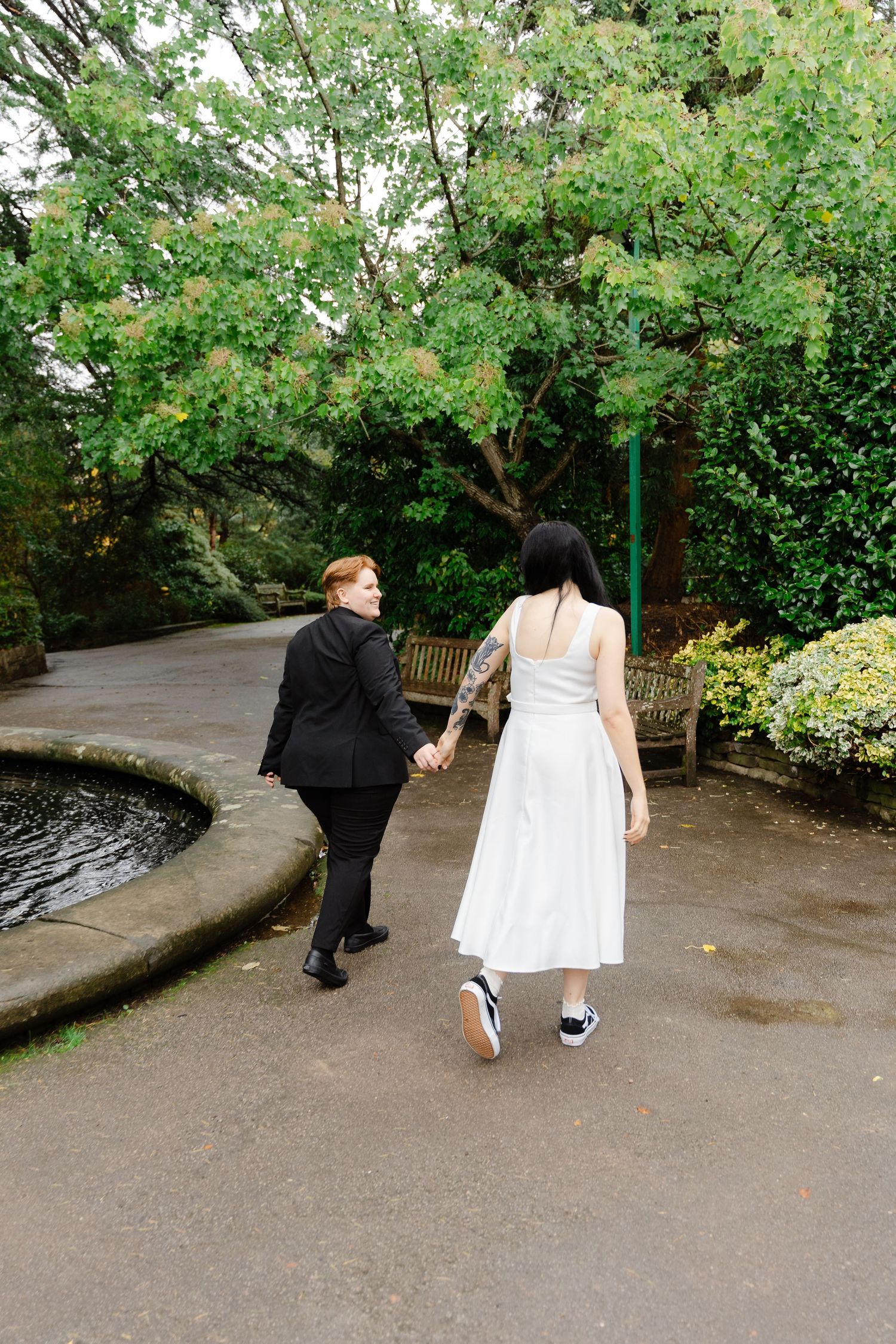 Two brides walk hand in hand through a lush garden, one in a black suit and one in a white dress.