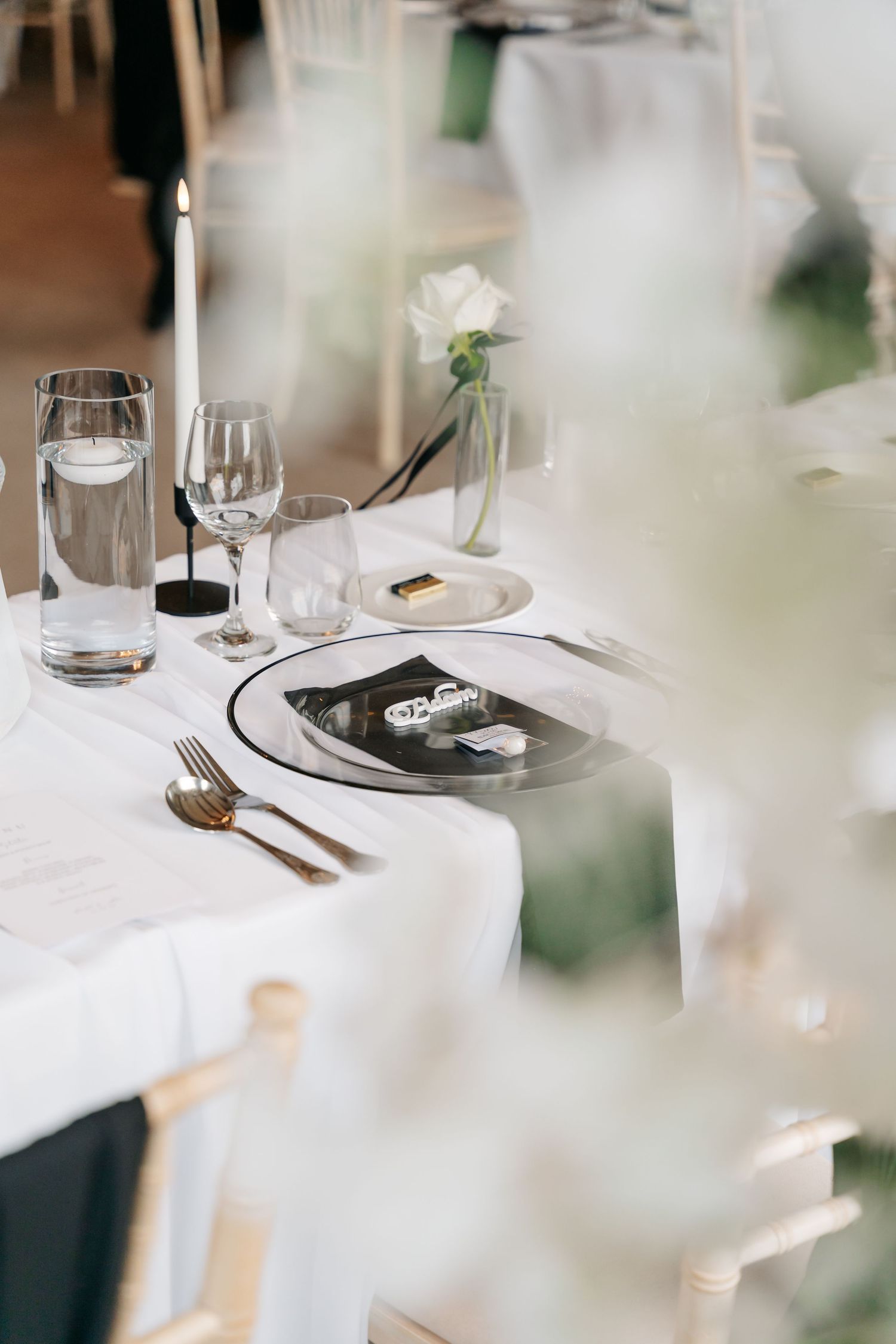 Elegant wedding table setting with white linen, gold cutlery, candle, glassware, and a white rose centerpiece.