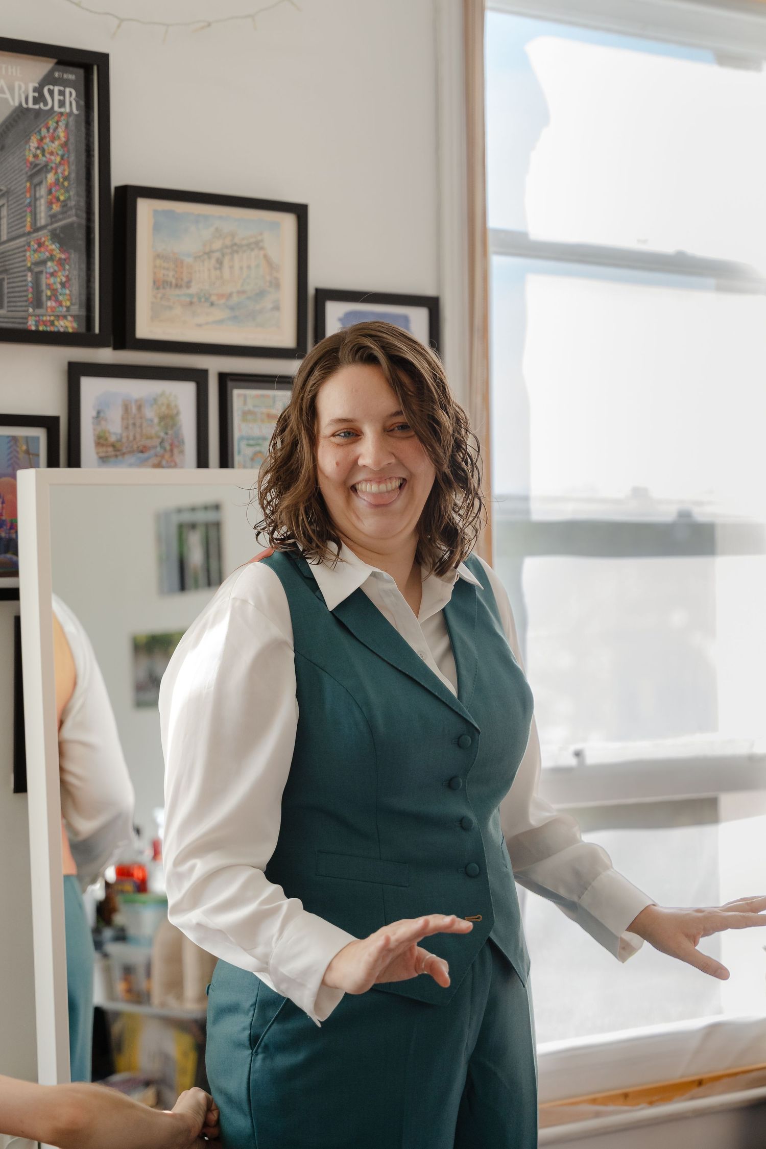 Smiling woman in green vest and white shirt stands in art-filled studio near window.
