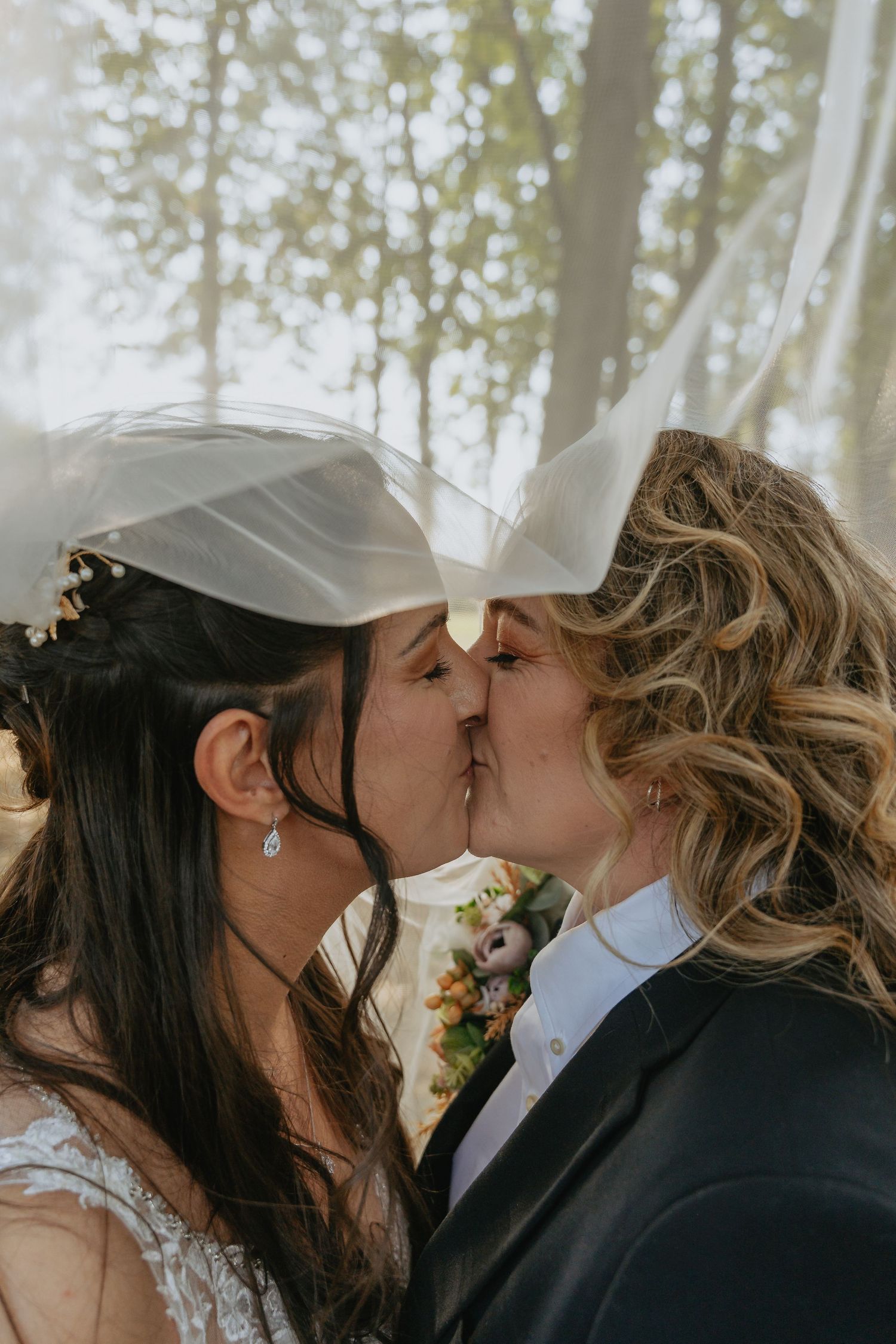 Two brides sharing a romantic kiss under a flowing veil on their wedding day outdoors.