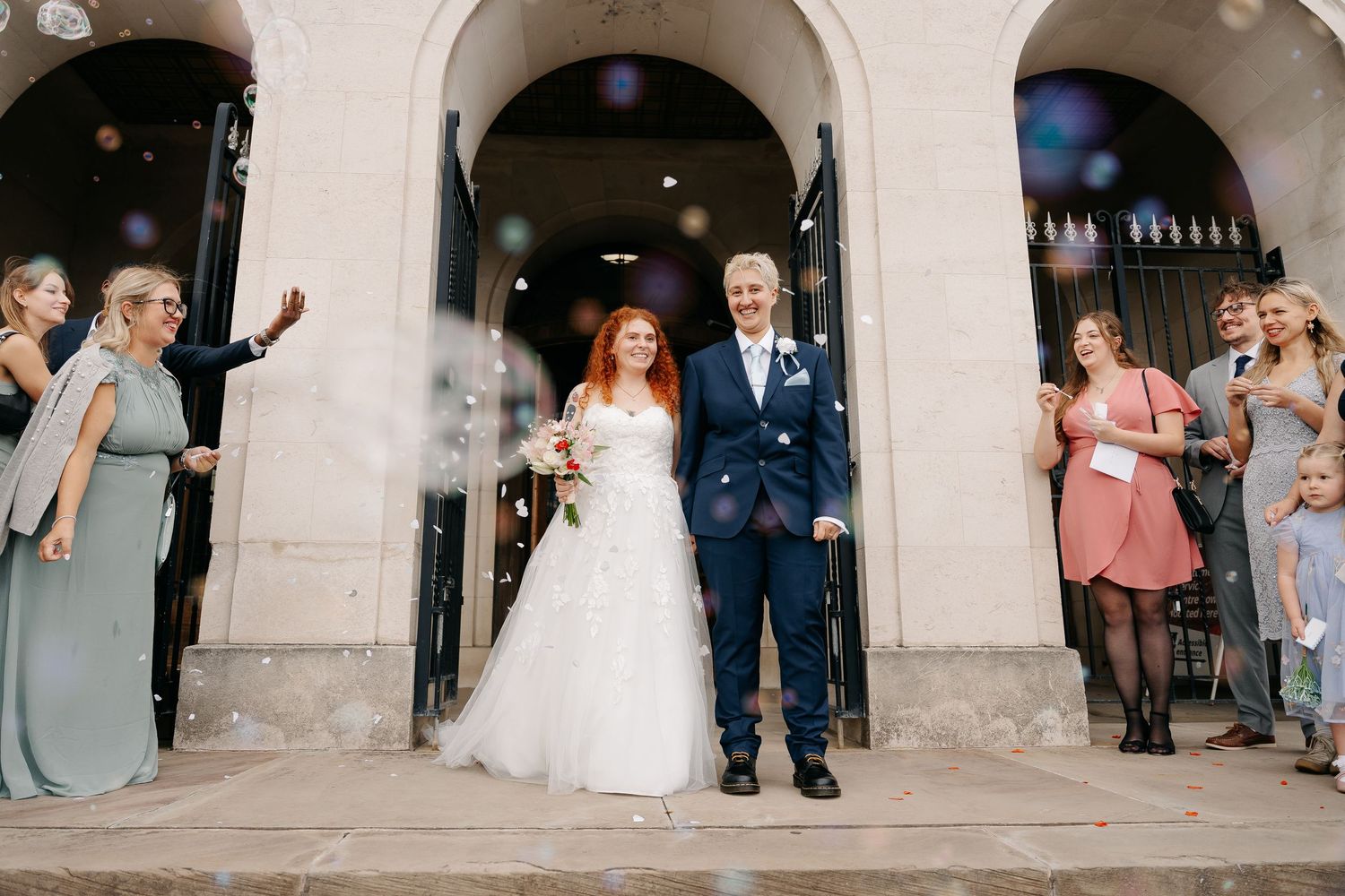 Bride and groom exit church doors as guests throw confetti, bride wearing white gown, groom in navy suit.