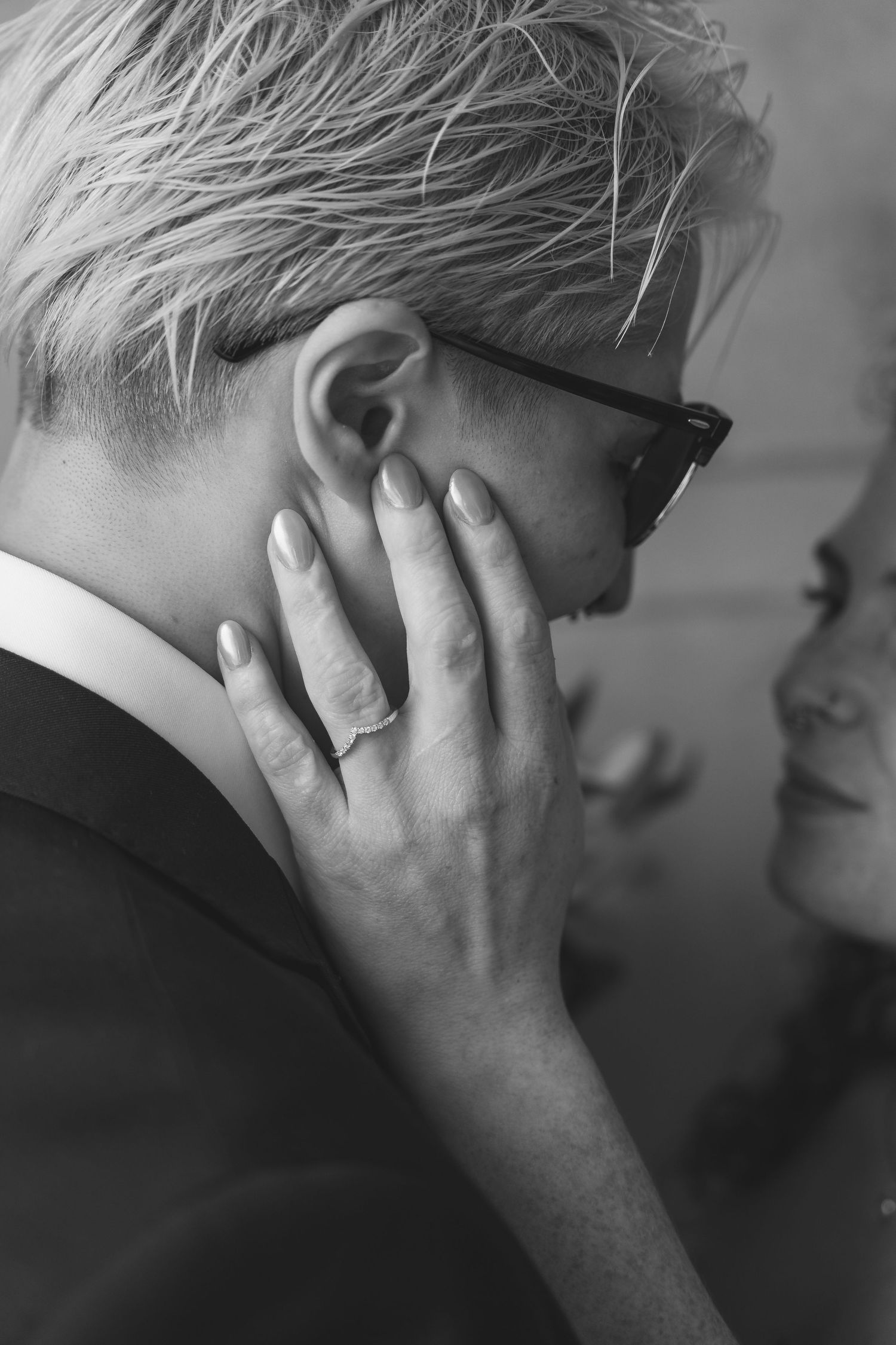 Black and white photo of a blonde person in glasses with a hand gently touching their neck, wearing a ring.