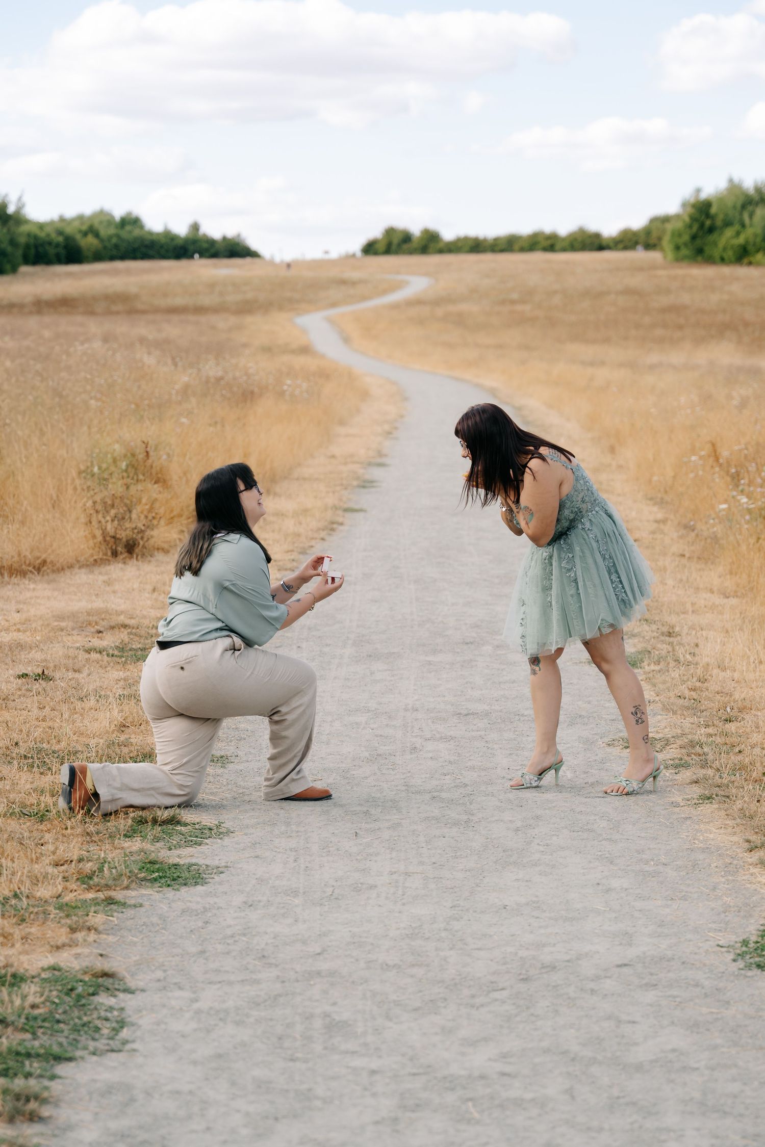 Person proposing on one knee to partner on scenic rural path, holding ring box, partner reacting with surprise in blue dress.