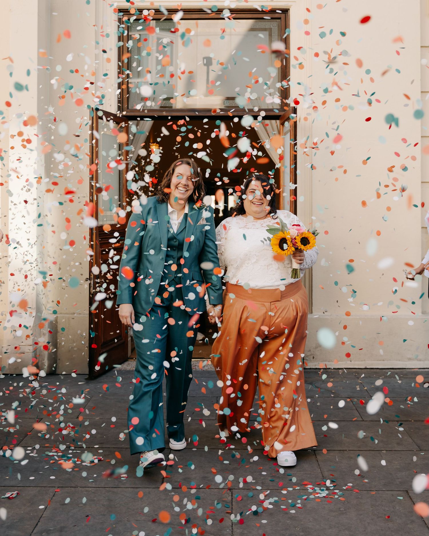 Two brides celebrate with colorful confetti shower outside venue, one in teal suit, one in white top and rust skirt.