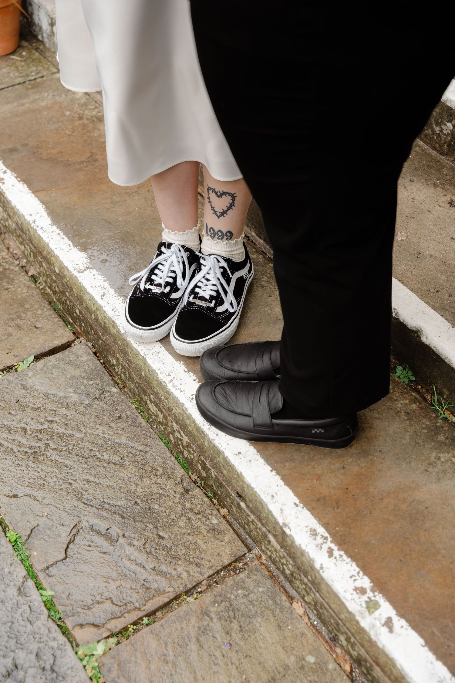 Close-up of couple's feet on stone steps, woman wearing black Vans sneakers with heart tattoo, man in black leather shoes.