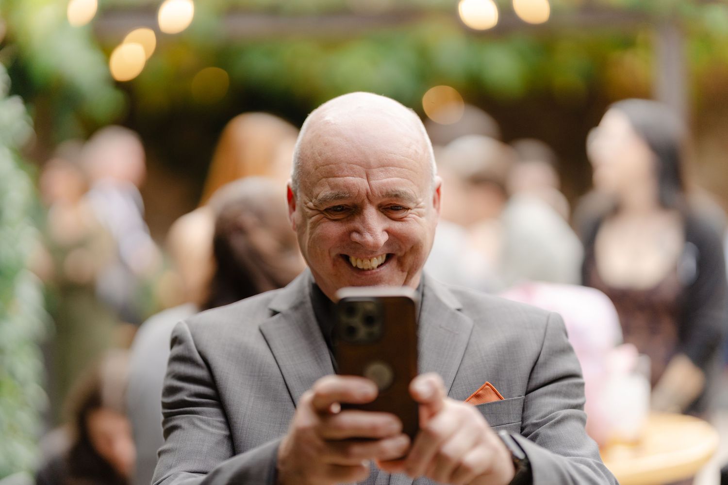 Smiling bald man in gray suit holding smartphone at outdoor event with bokeh lights.