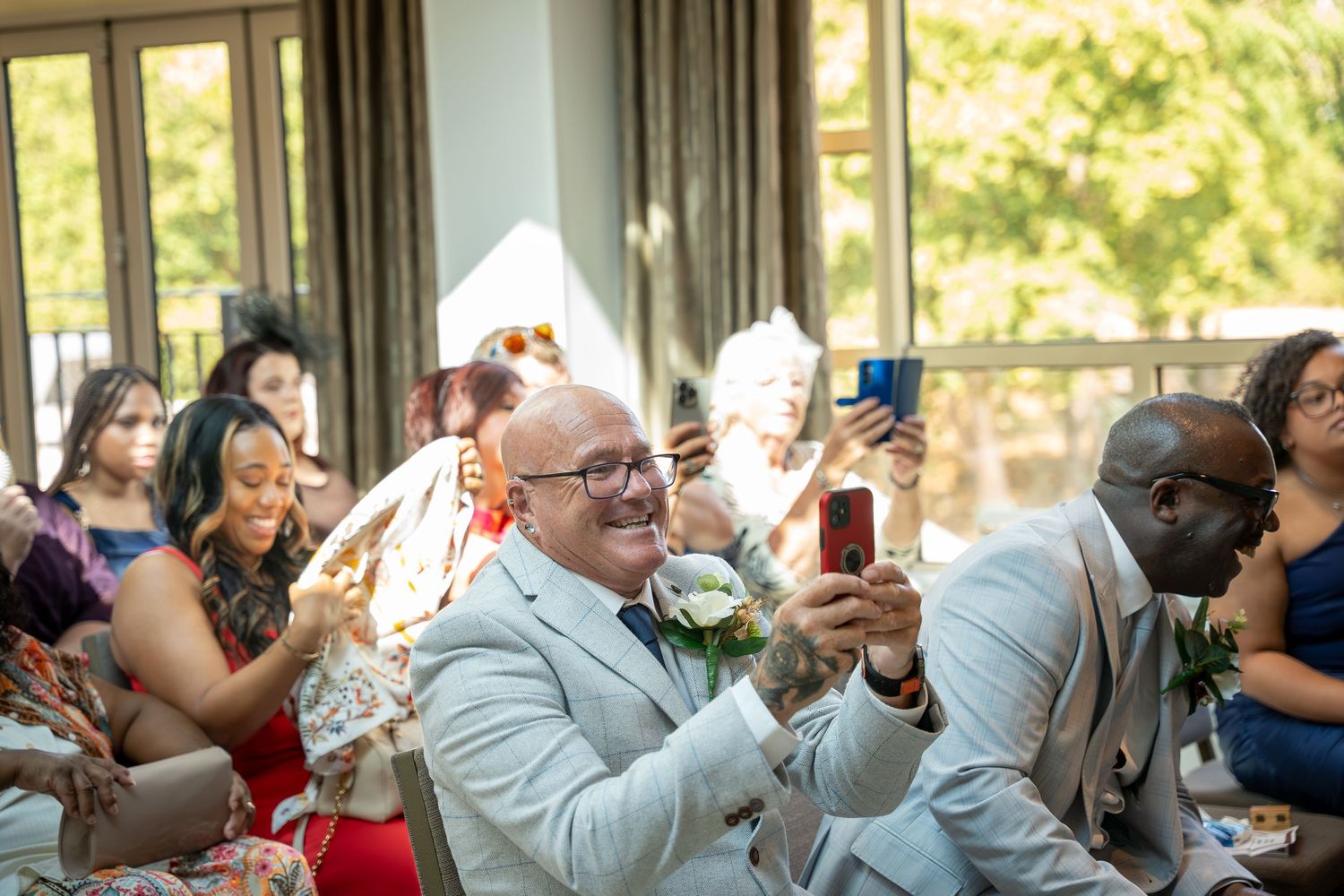 Guests smile and take photos on smartphones during a joyful wedding ceremony indoors with greenery visible outside.
