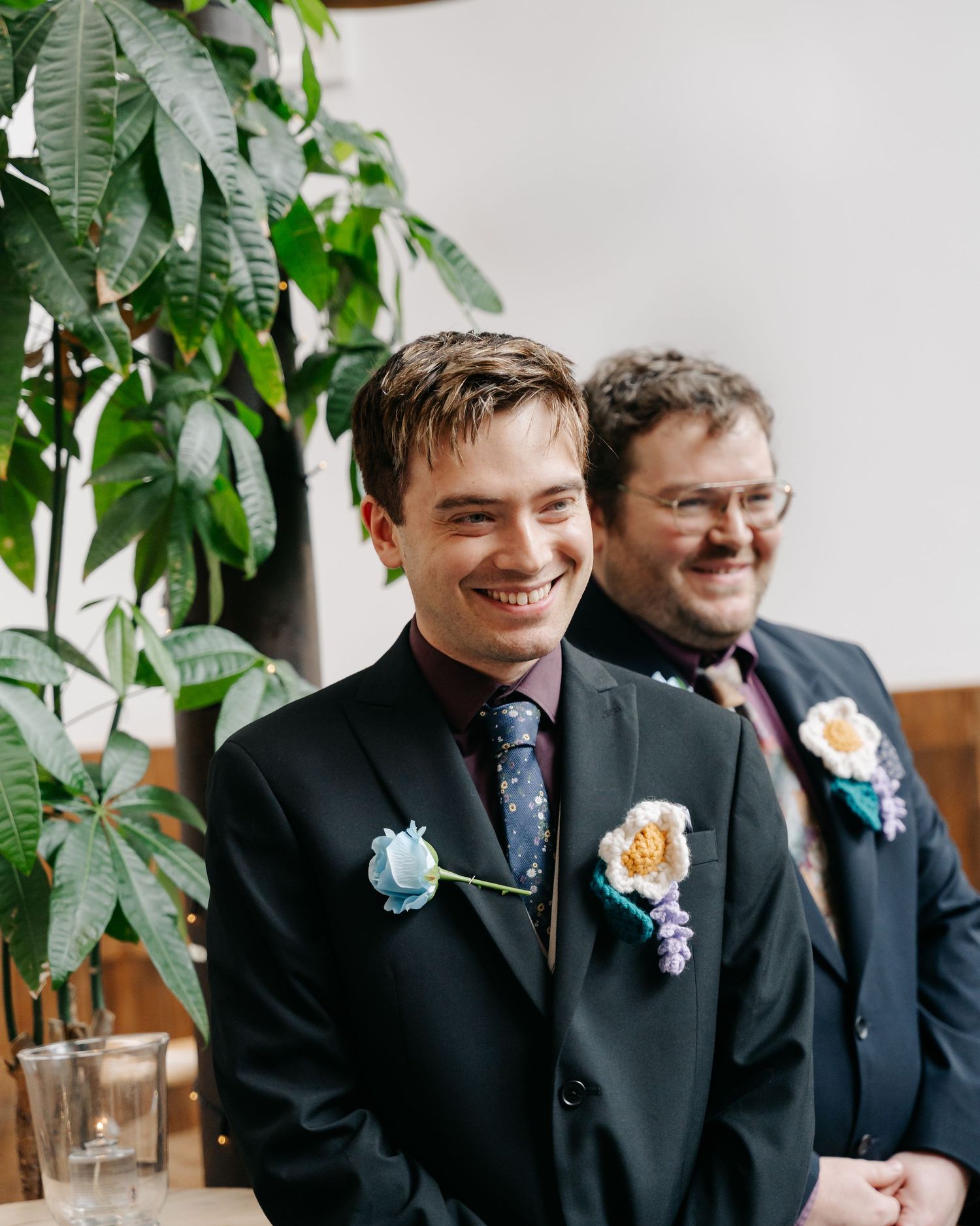 Two grooms smiling at wedding ceremony wearing dark suits with colorful floral boutonnieres, standing near lush green plants.