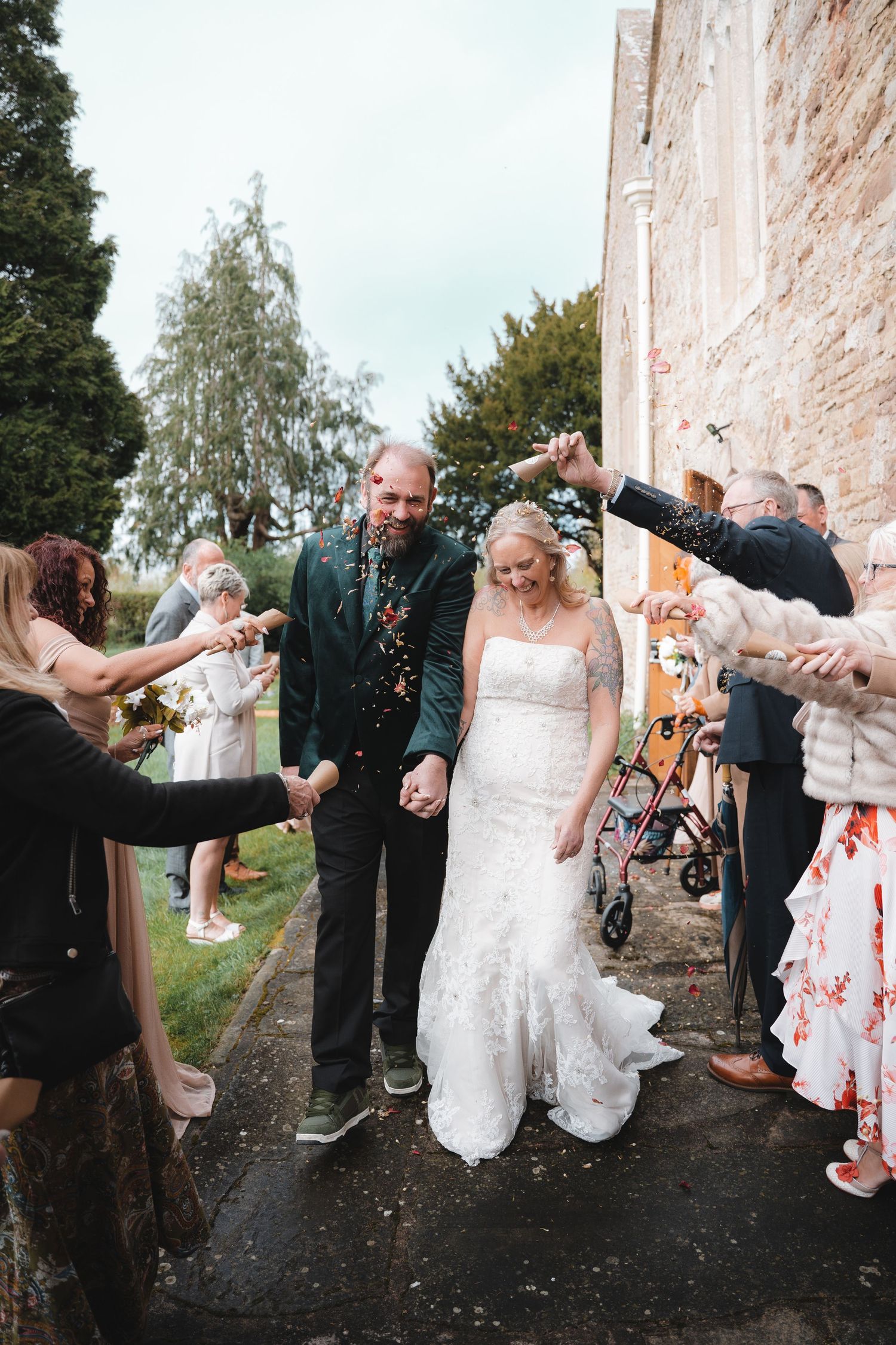 Bride and groom walk through confetti shower after wedding ceremony, guests lining path outside stone church.