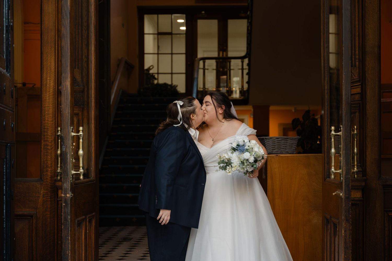 Bride and groom share a kiss in elegant wooden doorway, bride holding white bouquet in stunning off-shoulder wedding gown.