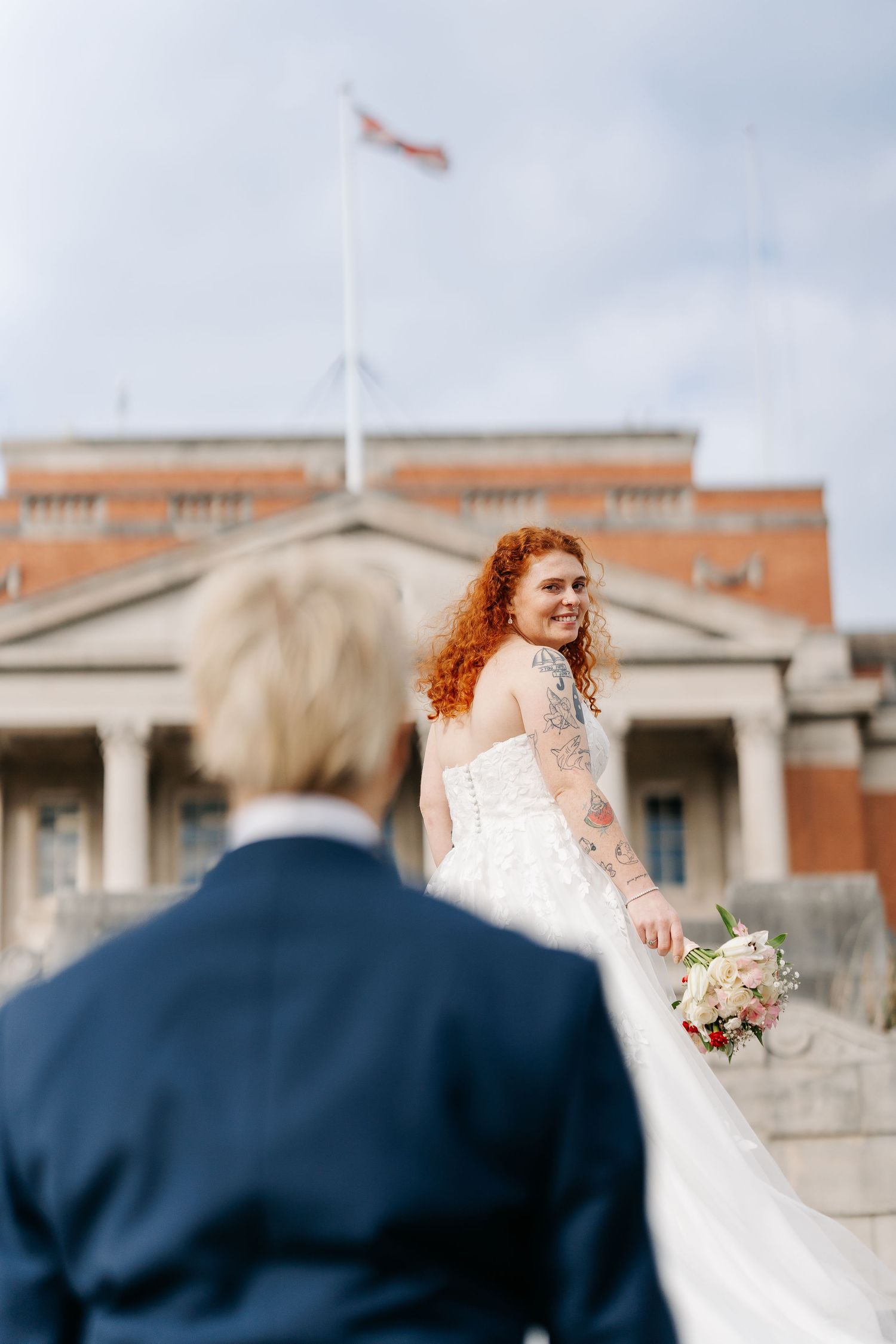 Red-haired bride in white gown holds bouquet on steps, smiling back at groom in navy suit before brick building.