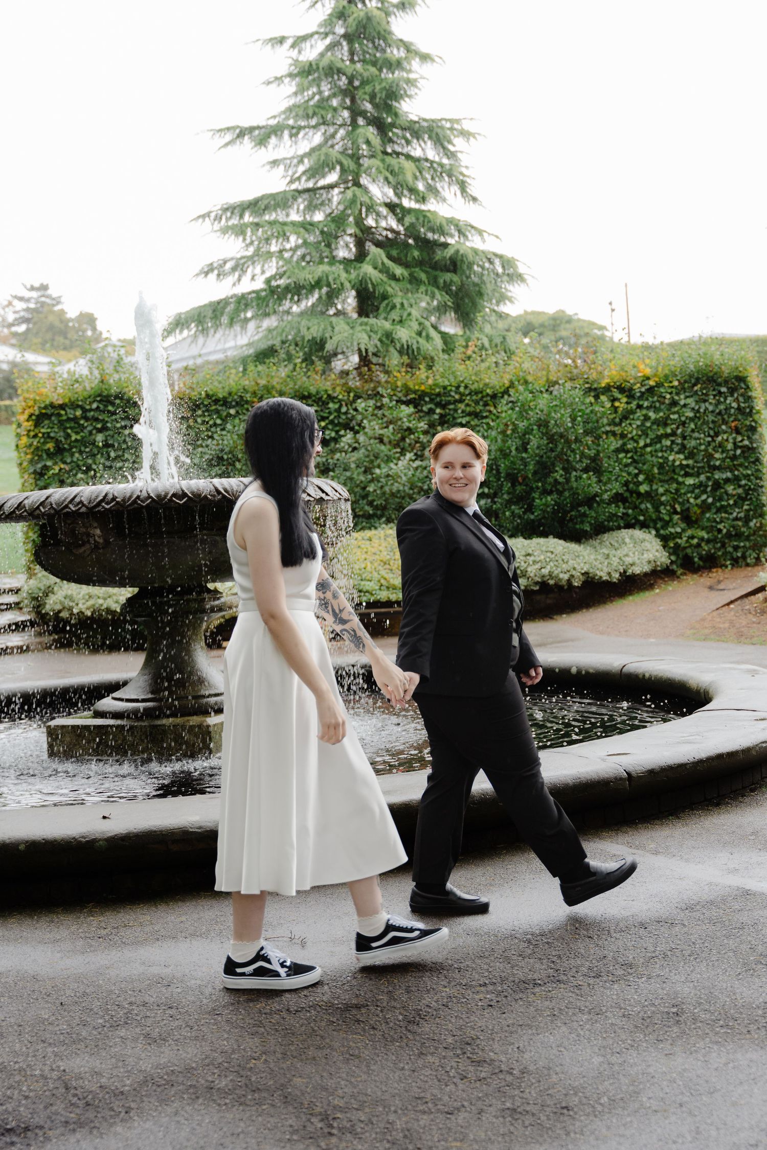 Two brides hold hands by a fountain in a garden, one in a white dress and Vans, the other in a black suit.