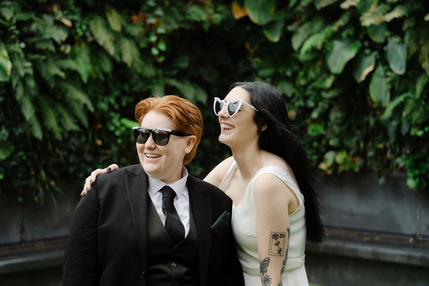 Two brides laughing together outdoors, one in a suit and one in a white dress, both wearing sunglasses before green foliage.