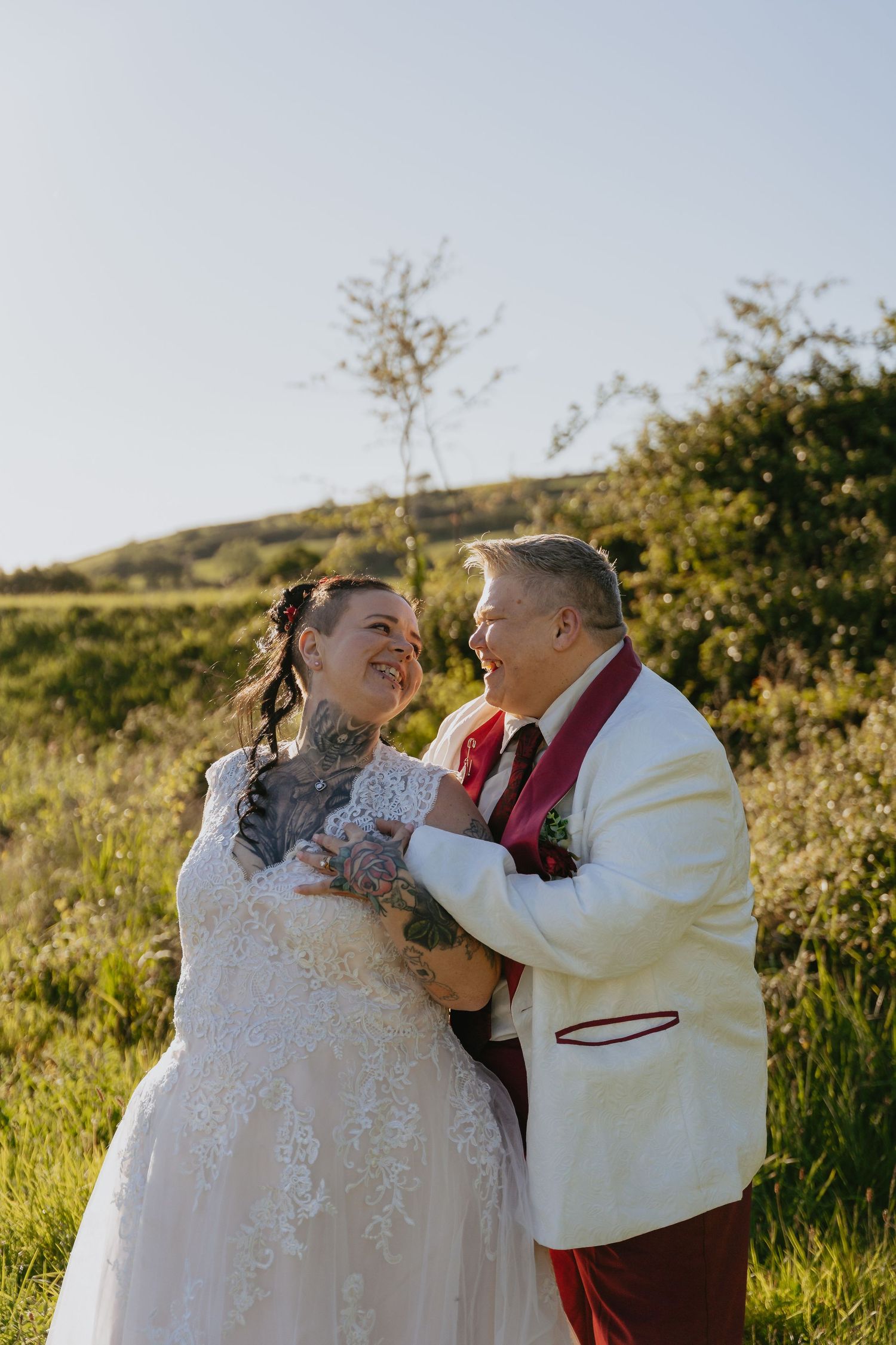 A laughing bride and groom in wedding attire share a joyful moment outdoors in a green field on their wedding day.
