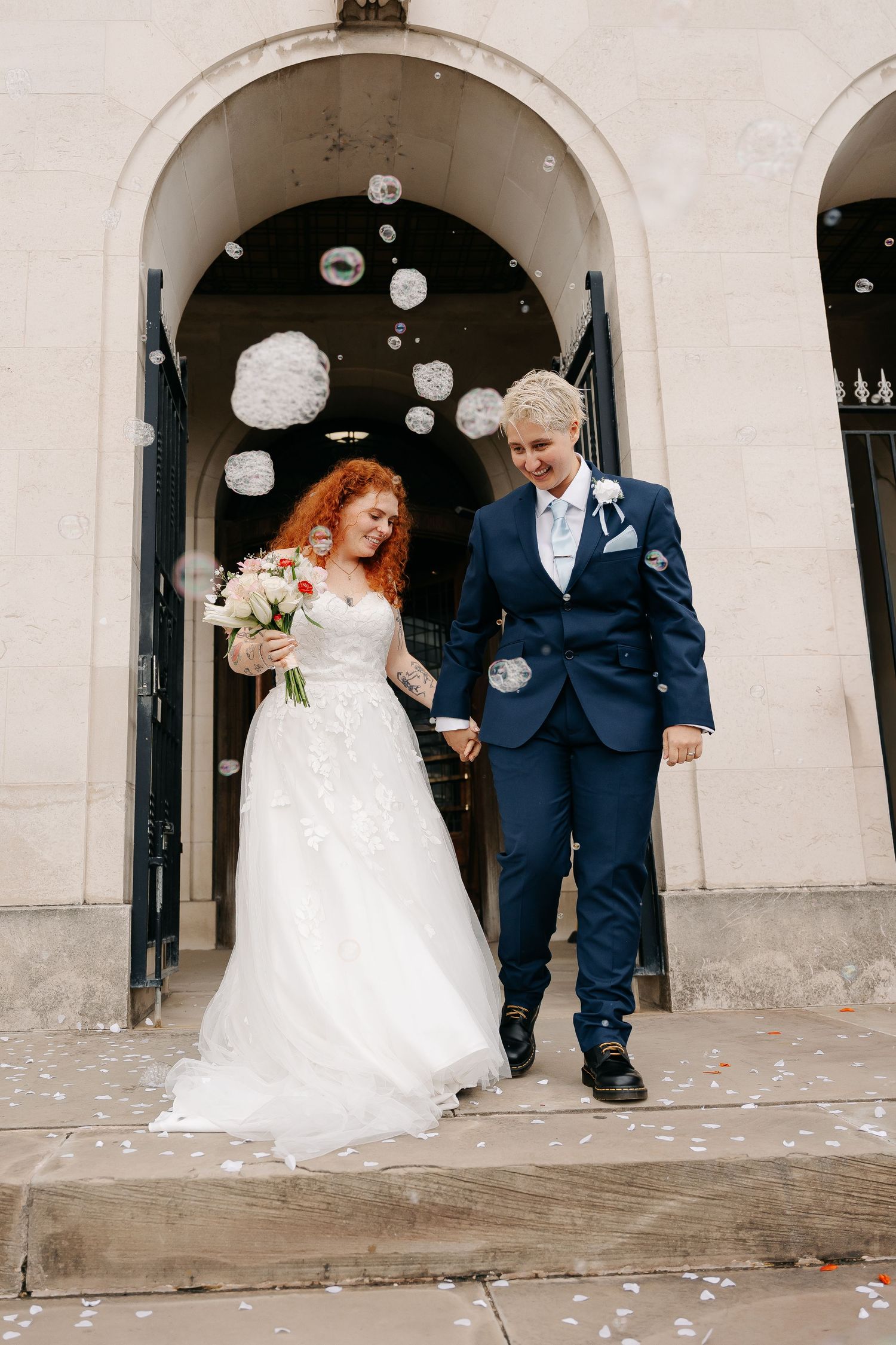 Two brides exit a church, one in a white gown holding flowers, the other in a navy suit, as bubbles float around them.