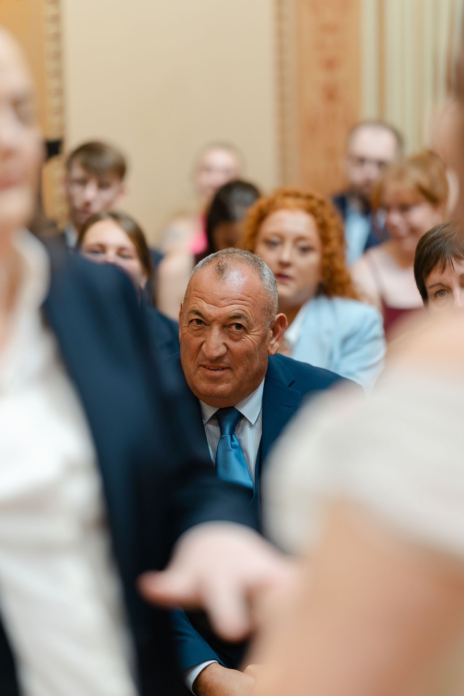 Man in blue suit and teal tie stands among wedding guests in a crowded ceremony hall.