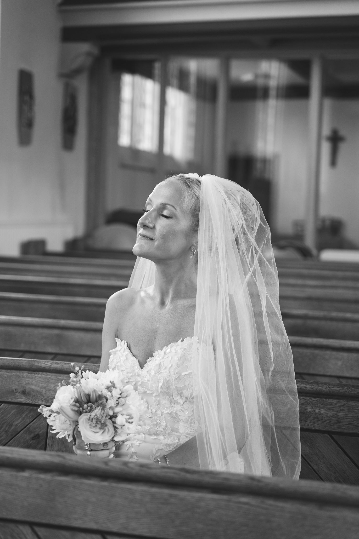 Bride in wedding dress and veil sits in church pew holding bouquet, eyes closed in peaceful prayer, black and white photo.