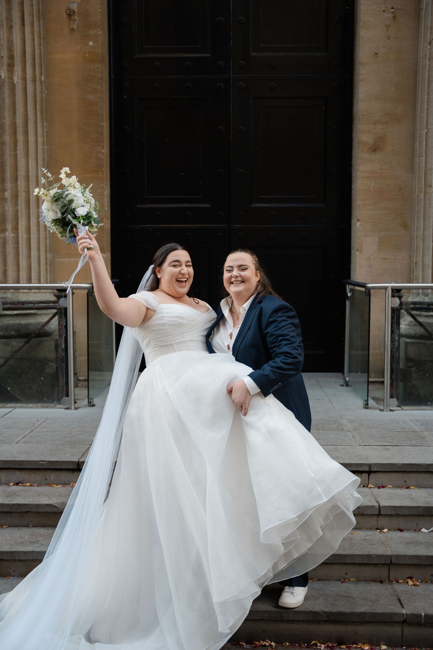Two brides celebrate joyfully on church steps, one in white gown with bouquet, other in navy suit.