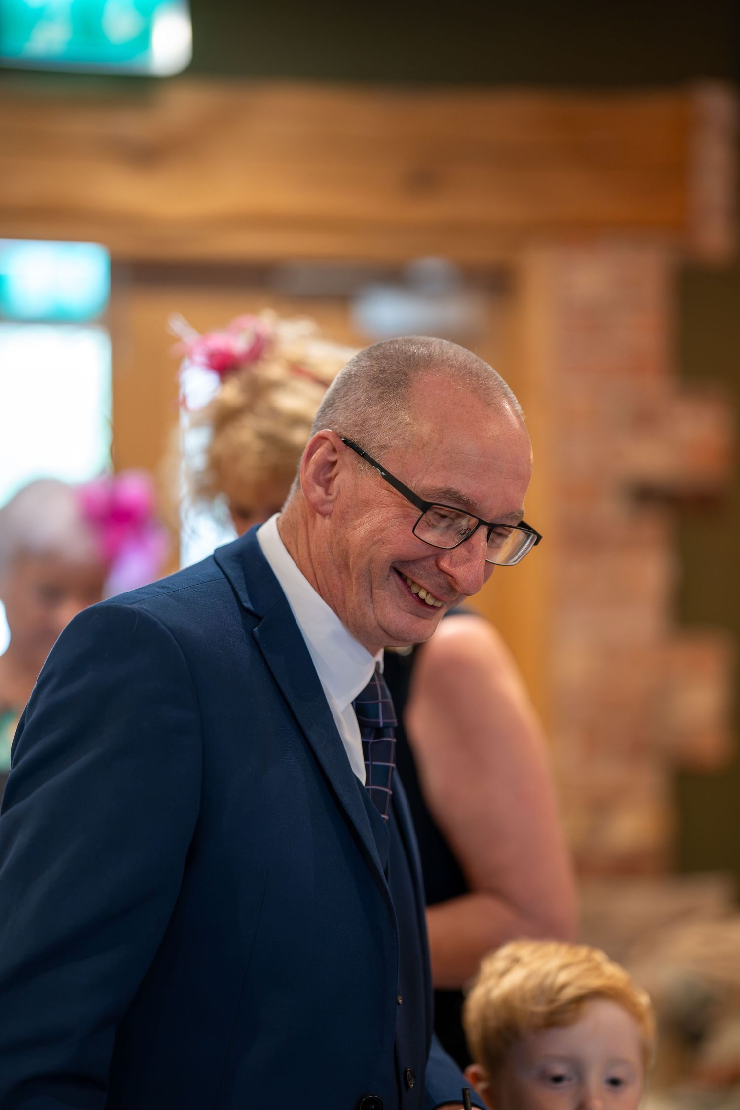 Smiling middle-aged man in navy blue suit and glasses at a formal event, with guests visible in background.