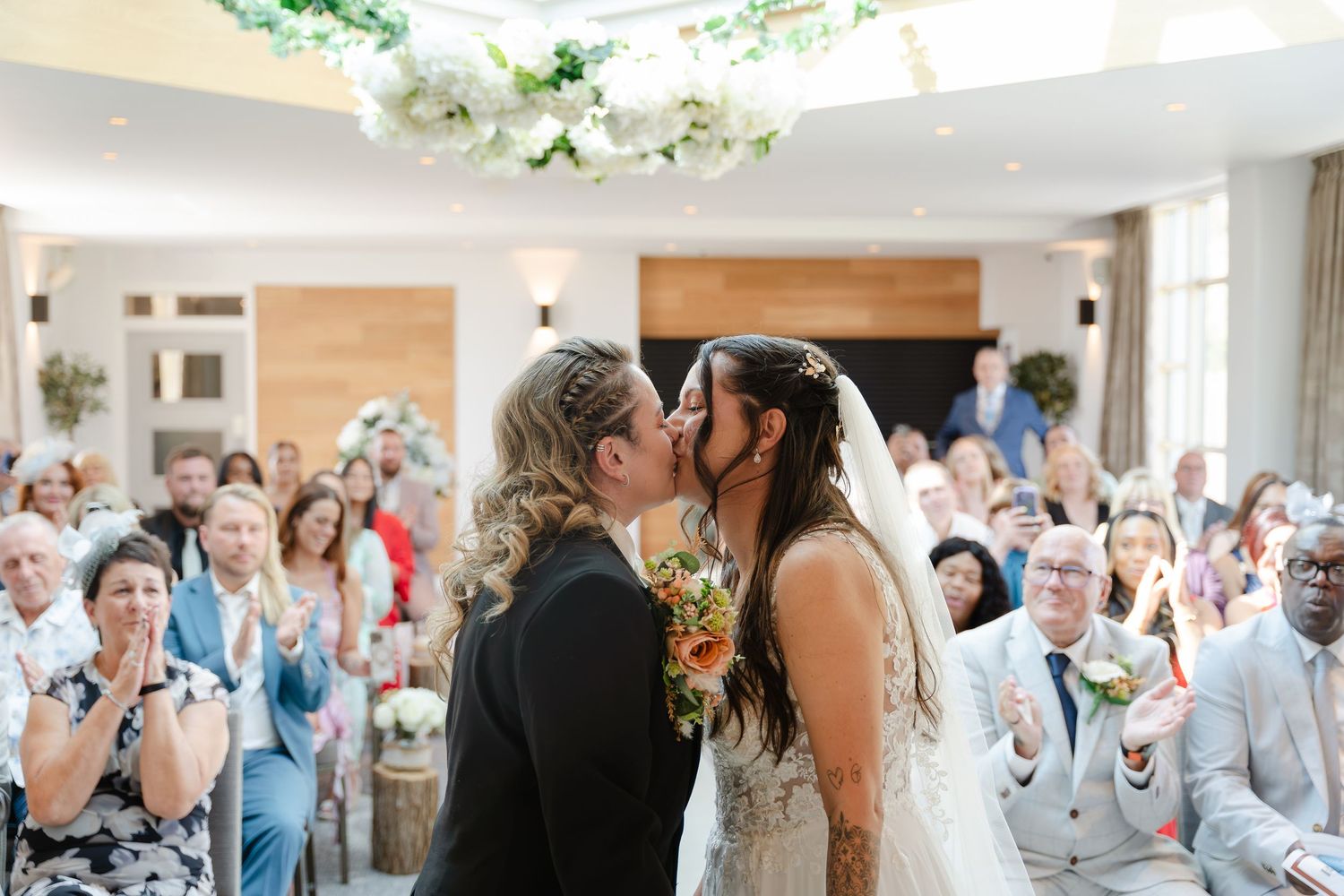 Two brides share their first kiss at a bright, elegant wedding ceremony surrounded by smiling guests and floral decorations.
