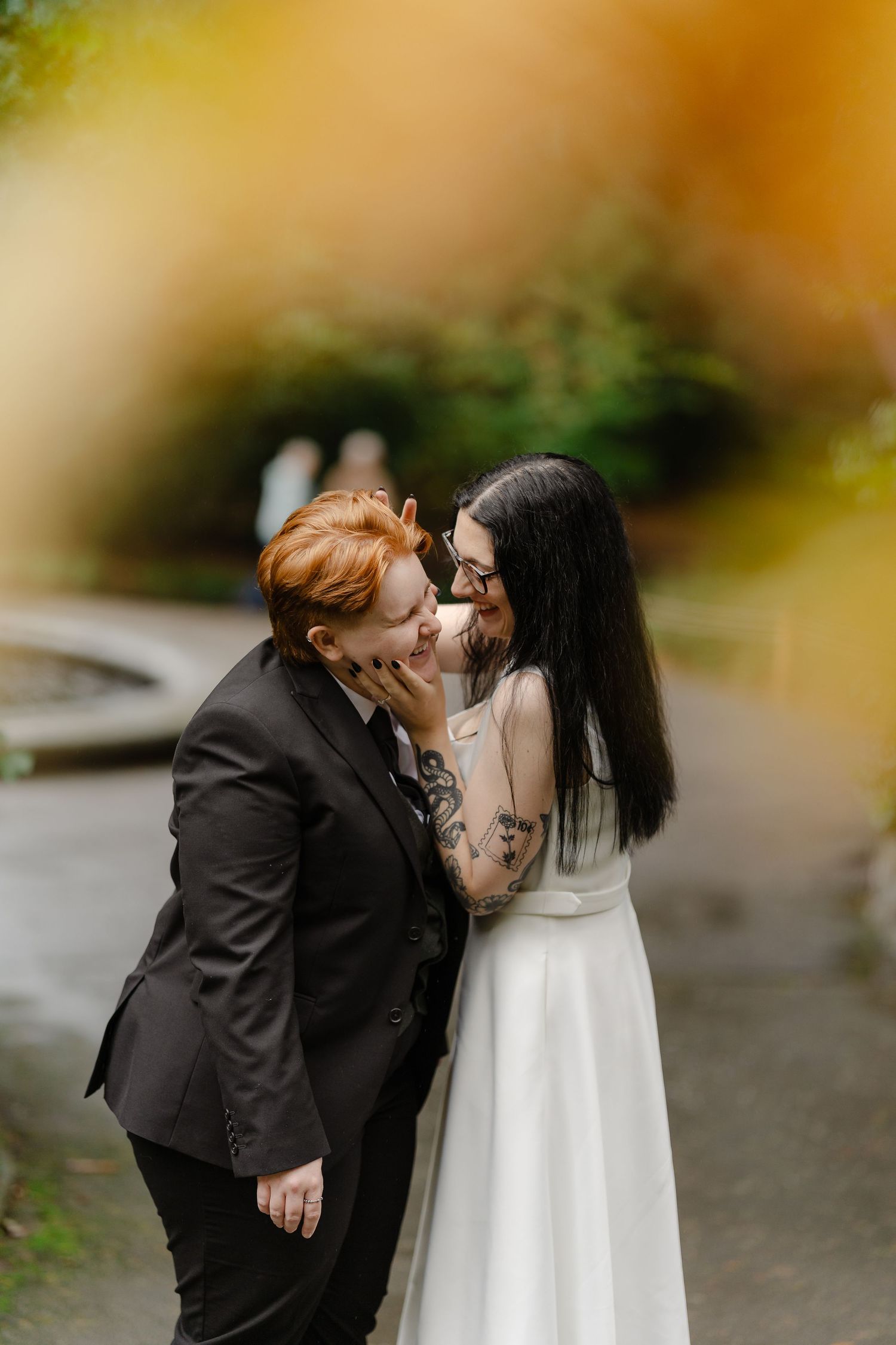 Two brides share a romantic moment, one in a black suit with red hair, the other in a white gown with dark hair and tattoos.