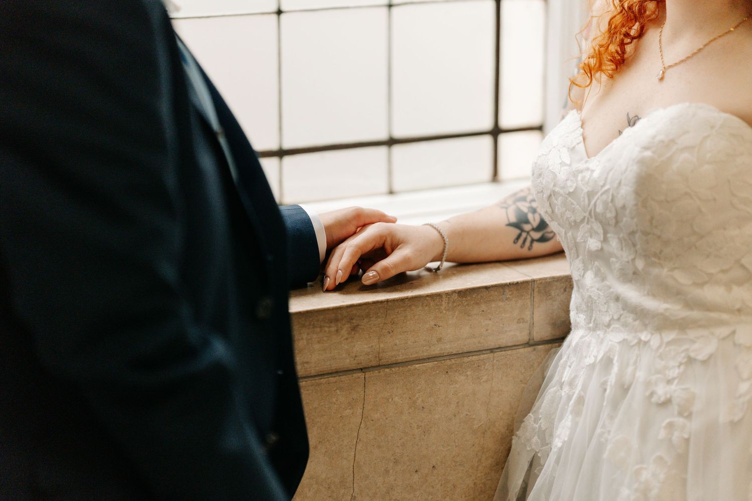 Bride in lace wedding dress with tattoo holds groom's hand on stone ledge during intimate wedding ceremony.