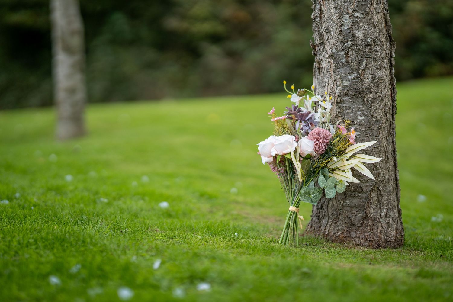 A rustic wedding bouquet with pink roses and wildflowers leaning against a tree trunk on a lush green lawn.