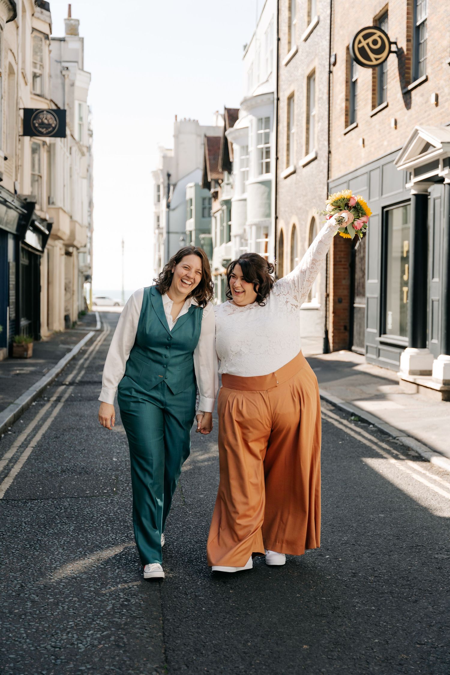 Two brides walk joyfully down a sunny street, one in a teal suit, the other in orange trousers holding a bouquet.