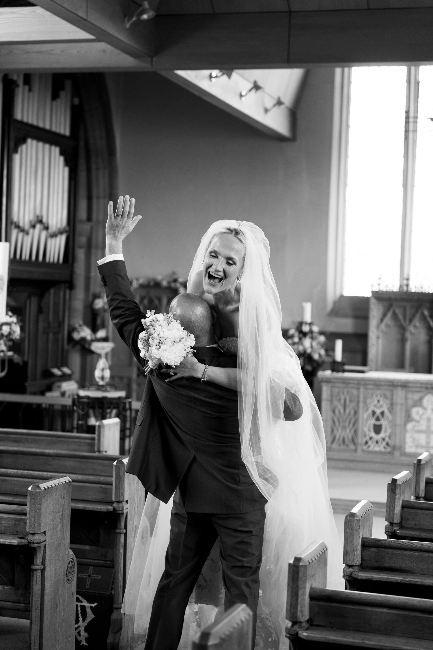 Black and white photo of joyful bride and groom celebrating in church aisle, bride holding bouquet and wearing veil.