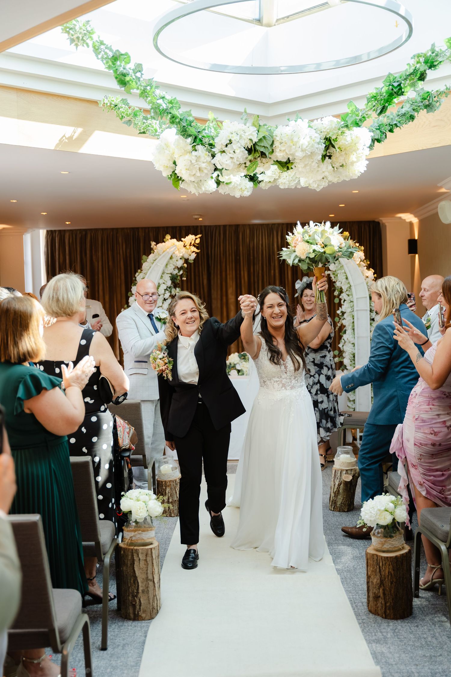 Two brides celebrate walking down the aisle after their wedding ceremony, surrounded by cheering guests and floral decor.