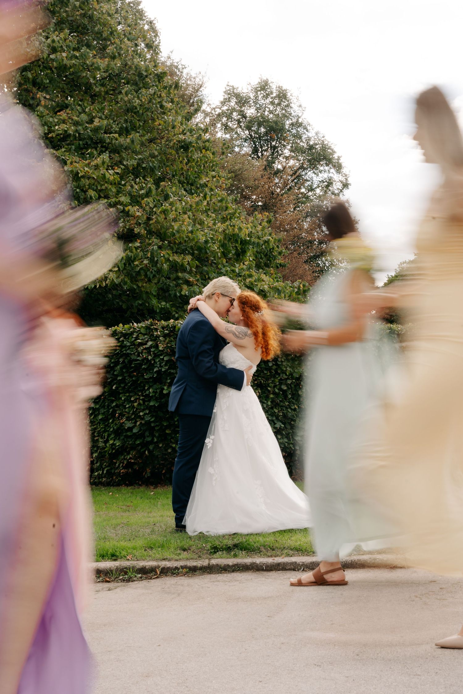 Bride and groom share a romantic kiss outdoors while blurred bridesmaids in colorful dresses walk past them.