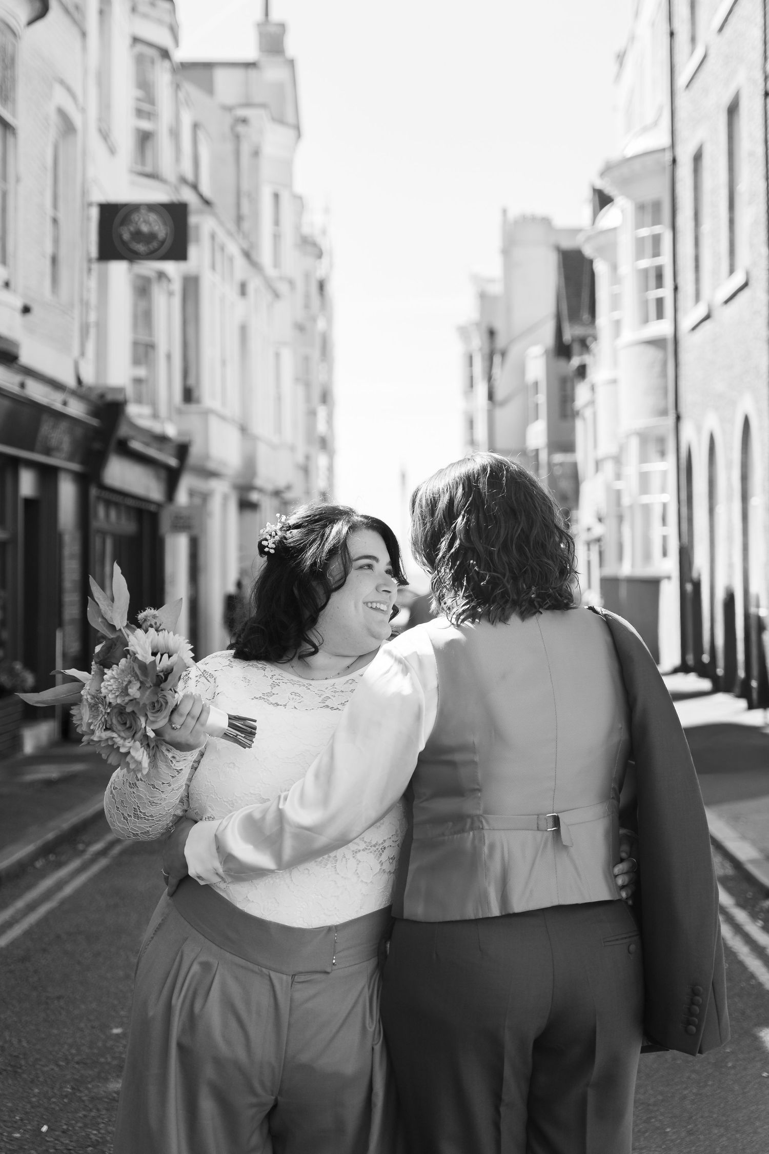 Black and white photo of a couple walking down a London street, bride holding bouquet, smiling at each other on wedding day.
