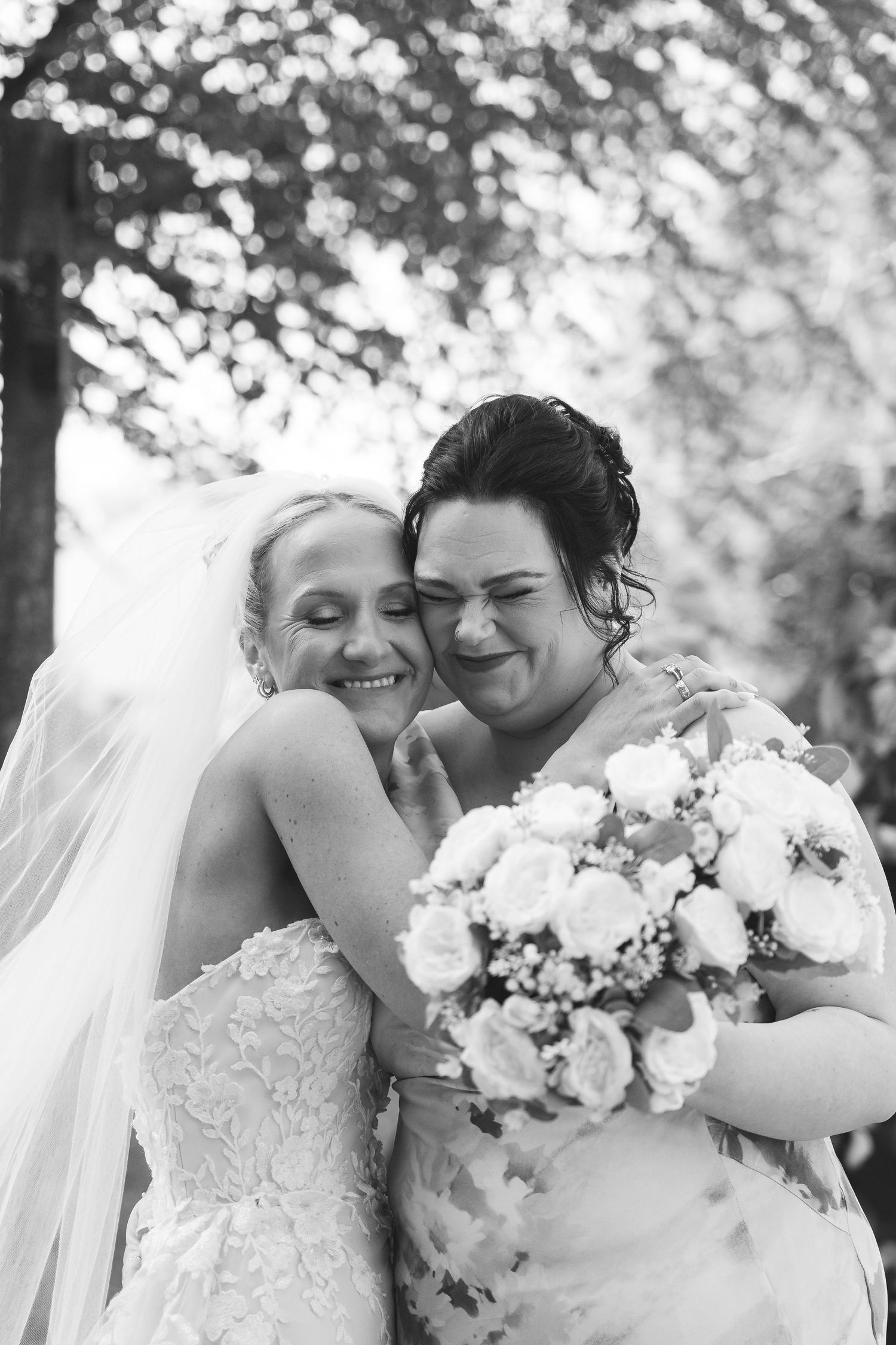 Two brides in white gowns embrace joyfully, holding a bouquet of roses, in a black and white outdoor wedding photo.