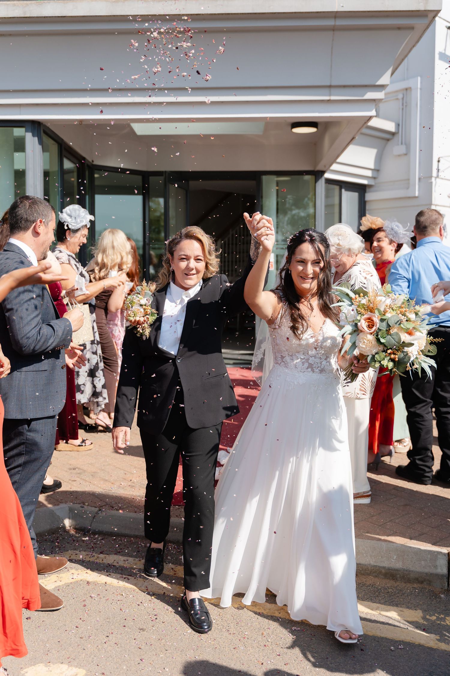 Two brides exit their wedding venue hand-in-hand, smiling as guests shower them with confetti.