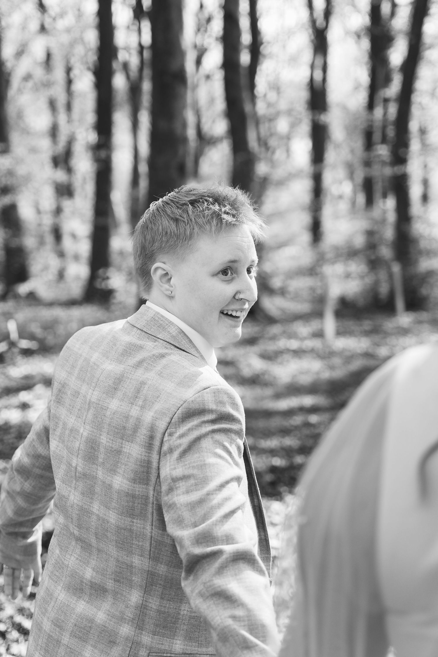 Black and white photo of a smiling person in a plaid suit jacket, standing in a wooded outdoor setting.