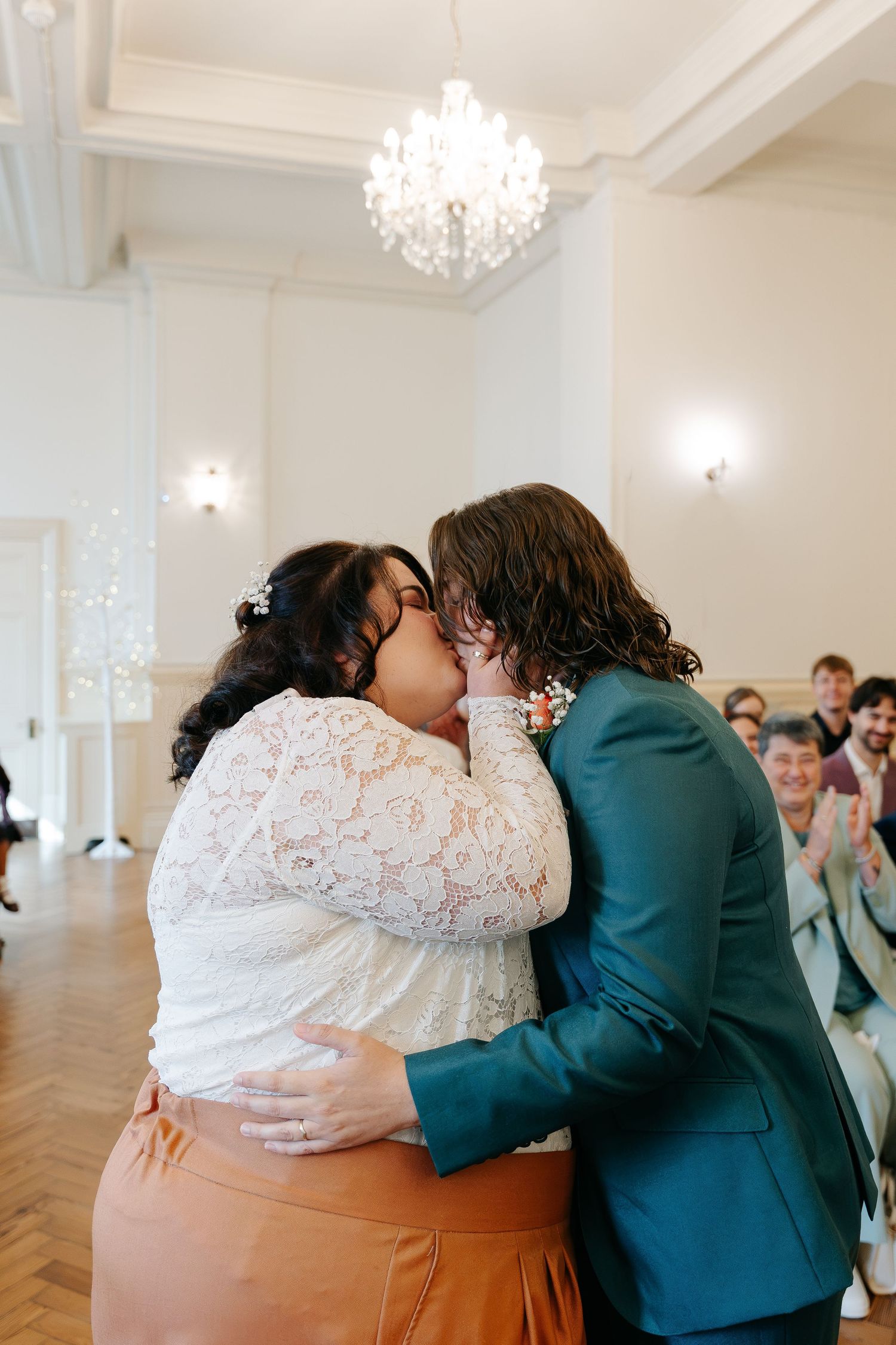 Two brides share their first kiss at wedding ceremony in elegant white ballroom with chandelier.