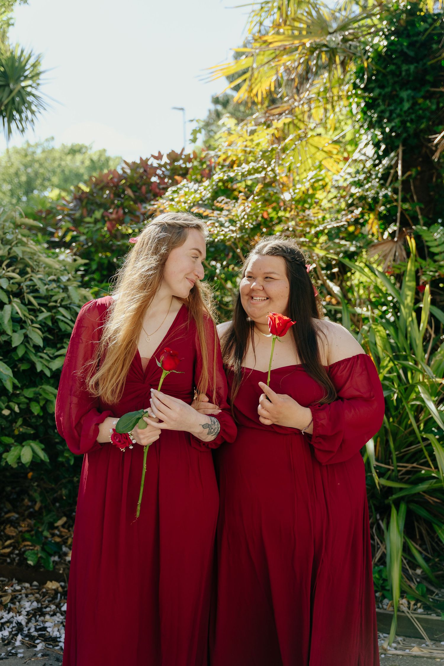 Two women in matching red dresses holding red roses smile at each other in a lush garden setting.